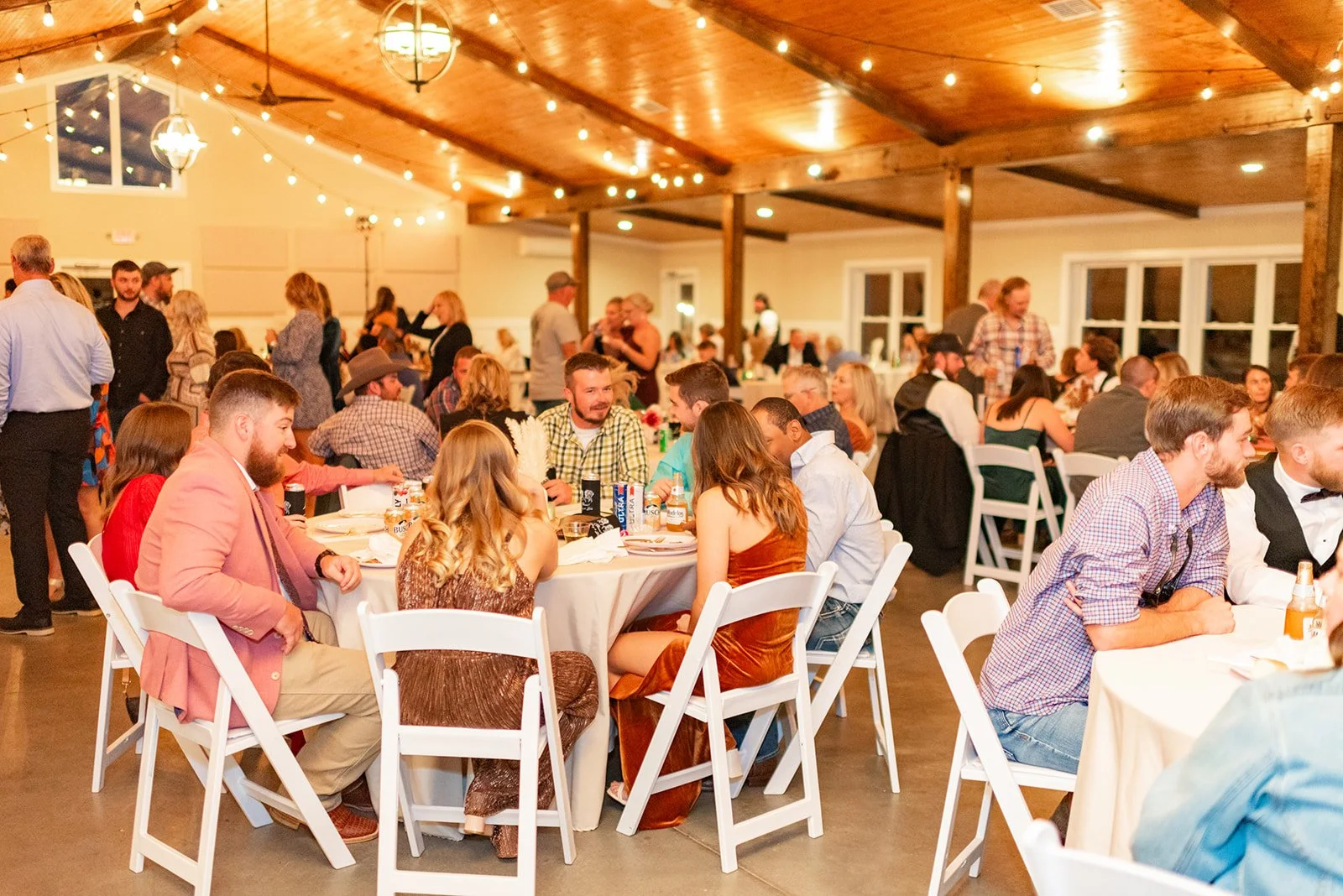 Guests seated at round tables with white chairs at a wedding reception, enjoying food and drinks in a decorated banquet hall with string lights and a wooden ceiling.