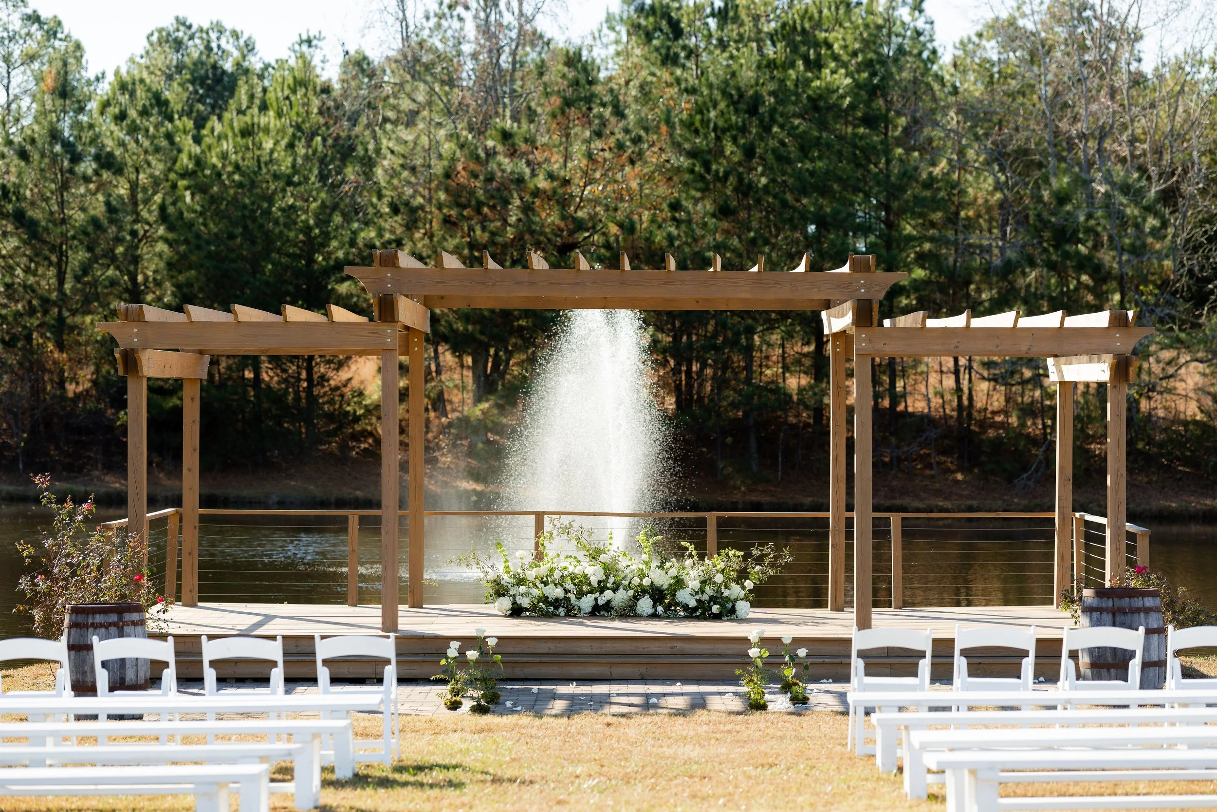 Wooden outdoor arch structure with water fountain behind it, decorated with white flowers at the base, set on a platform near a body of water, surrounded by trees.