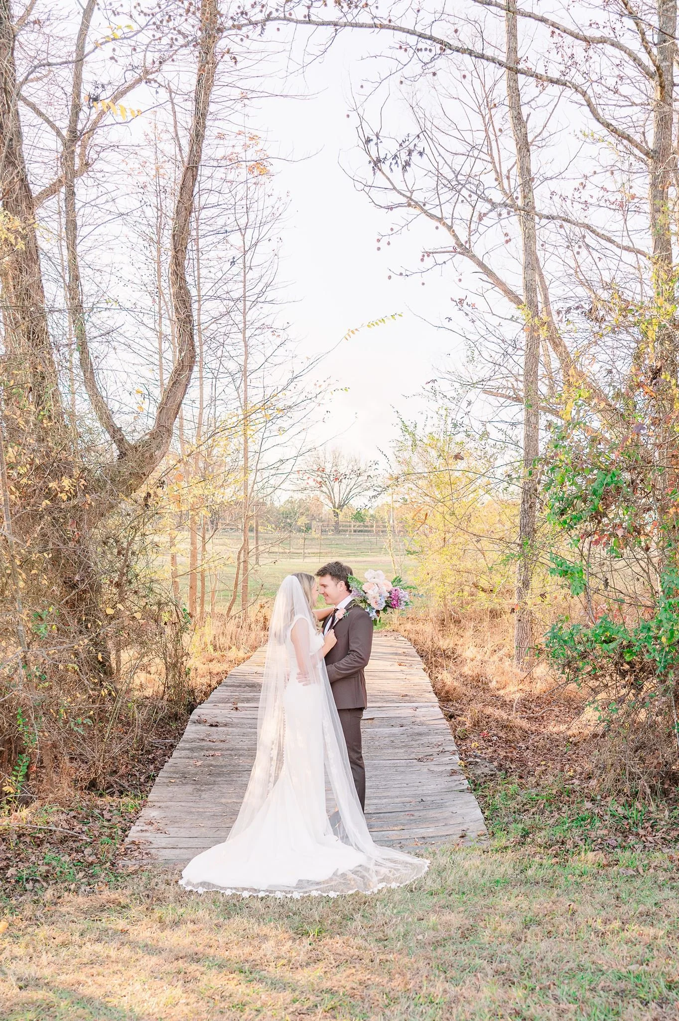 A bride and groom stand close together on a wooden bridge in a wooded area during autumn, gazing into each other's eyes, with the bride holding a bouquet of flowers.