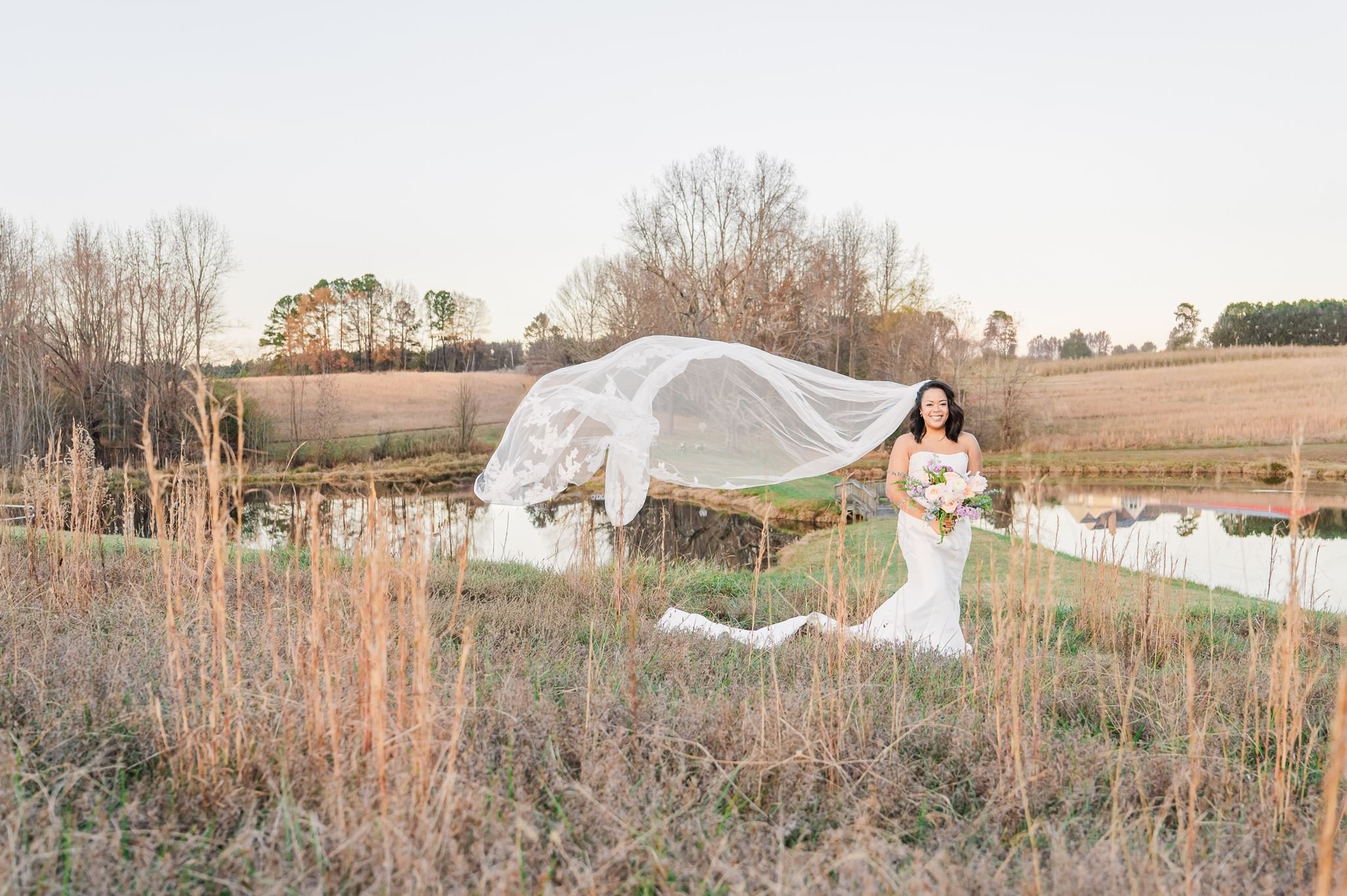 A woman in a white wedding dress holding a bouquet stands outdoors near a small lake, with leafless trees and a hilly landscape in the background during late fall or winter.