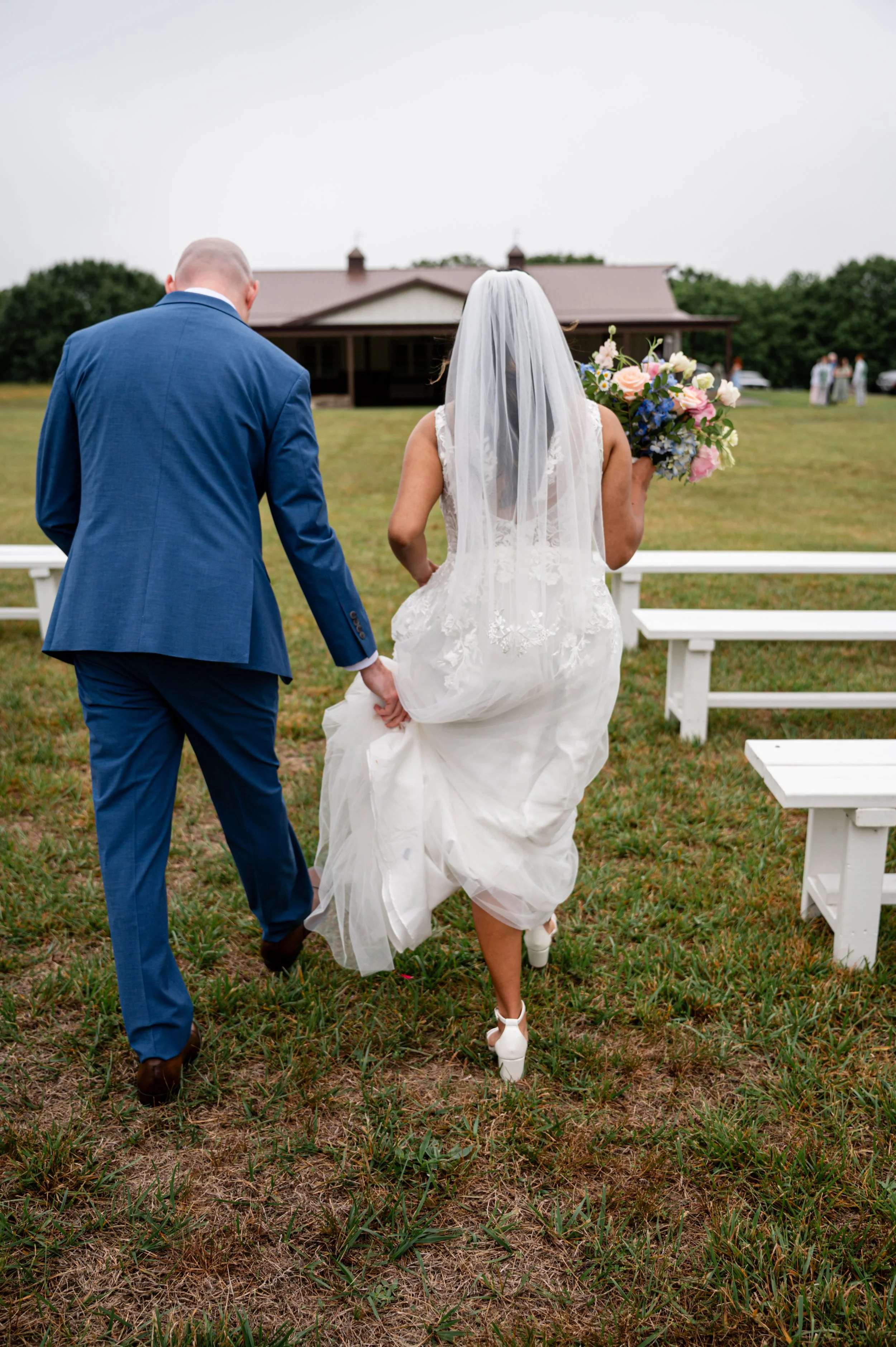 A bride and groom walking away from the camera in a grassy outdoor setting, with a house in the background, during a wedding ceremony. The bride is holding a bouquet of flowers and wears a white lace wedding dress with a veil. The groom is in a blue 