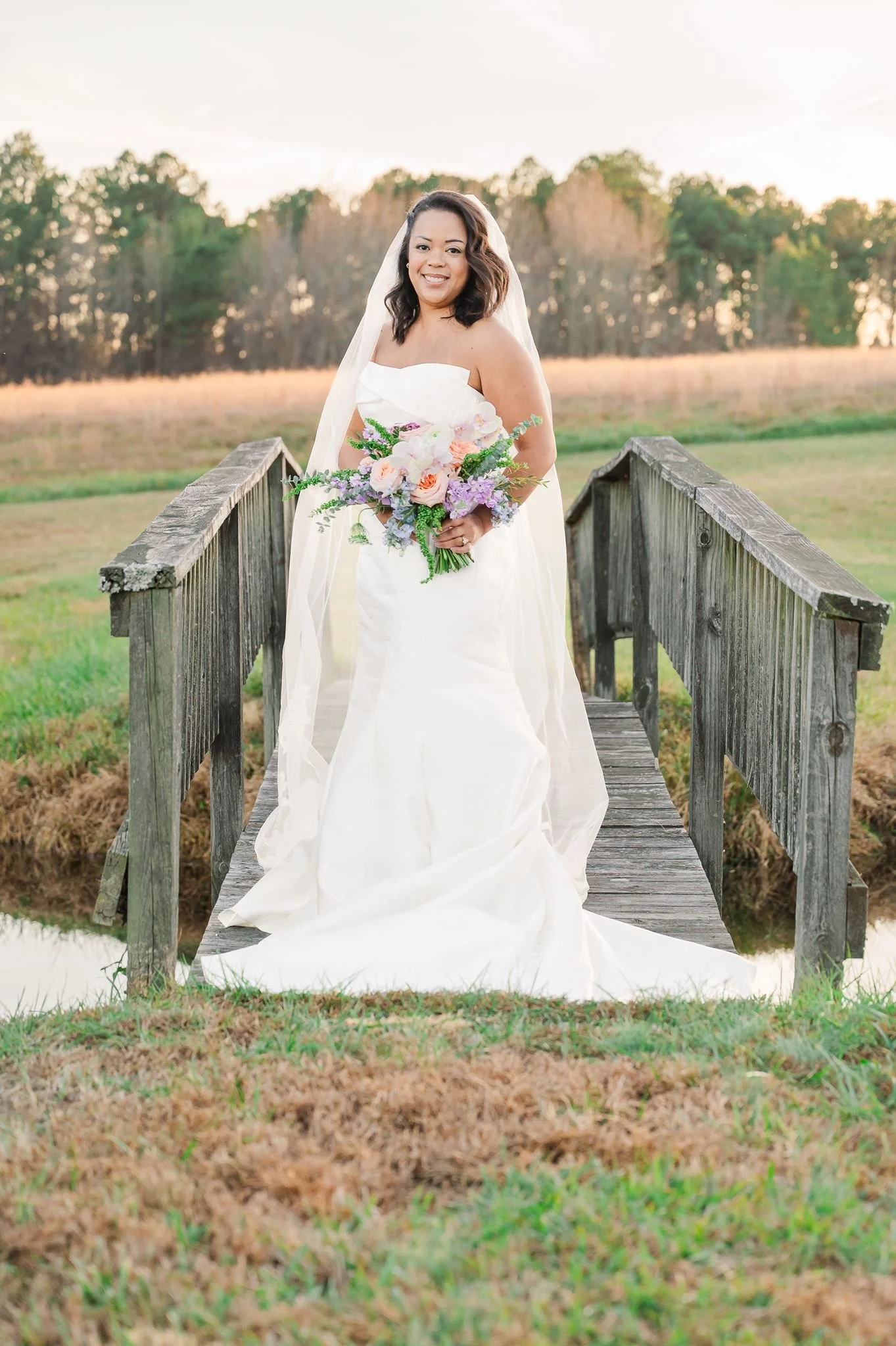 A smiling bride in a white wedding dress holding a colorful bouquet standing on a small wooden bridge outdoors at sunset.