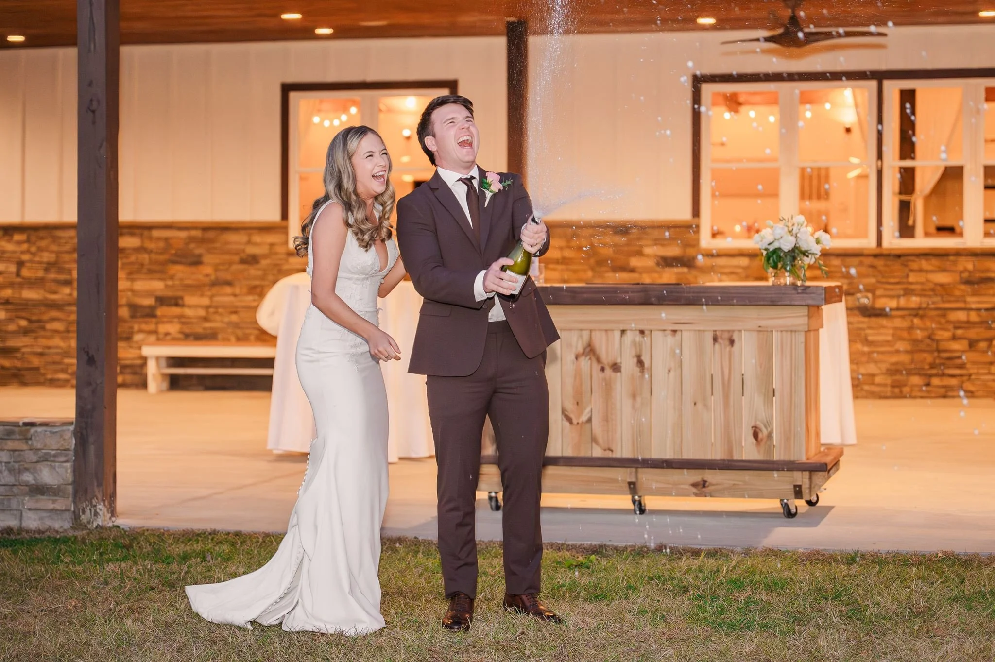 A bride and groom celebrating their wedding outdoors, with the groom opening a bottle of sparkling wine, causing it to spray as they laugh and enjoy the moment.