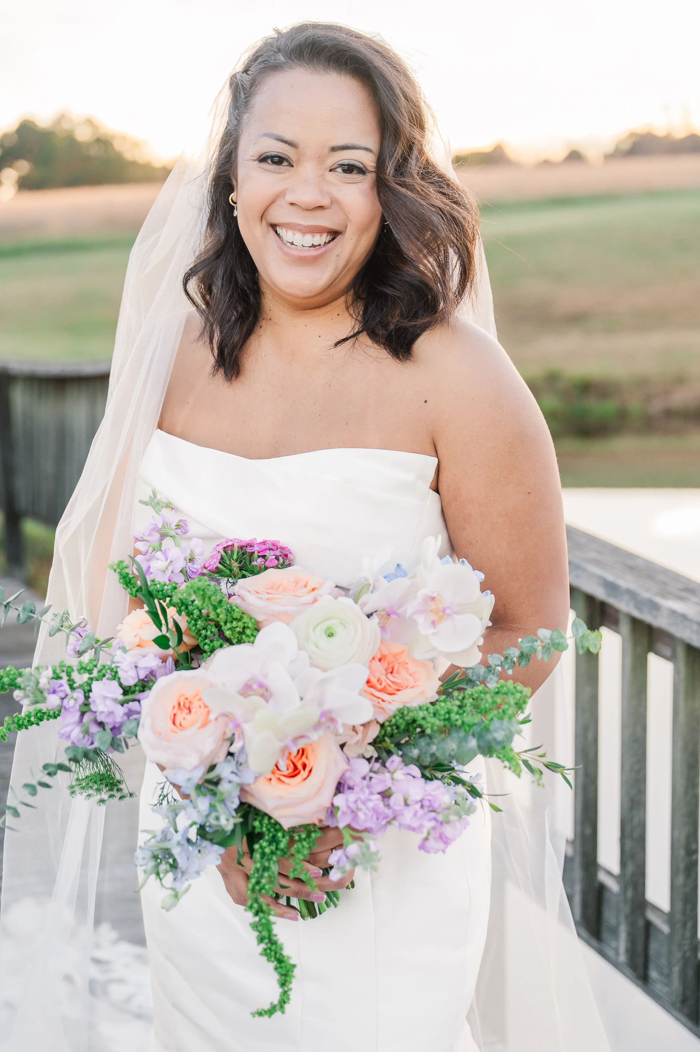 A bride in a wedding dress holding a large bouquet of colorful flowers by Stem and Petal, smiling on our bridge at Sable and Vine