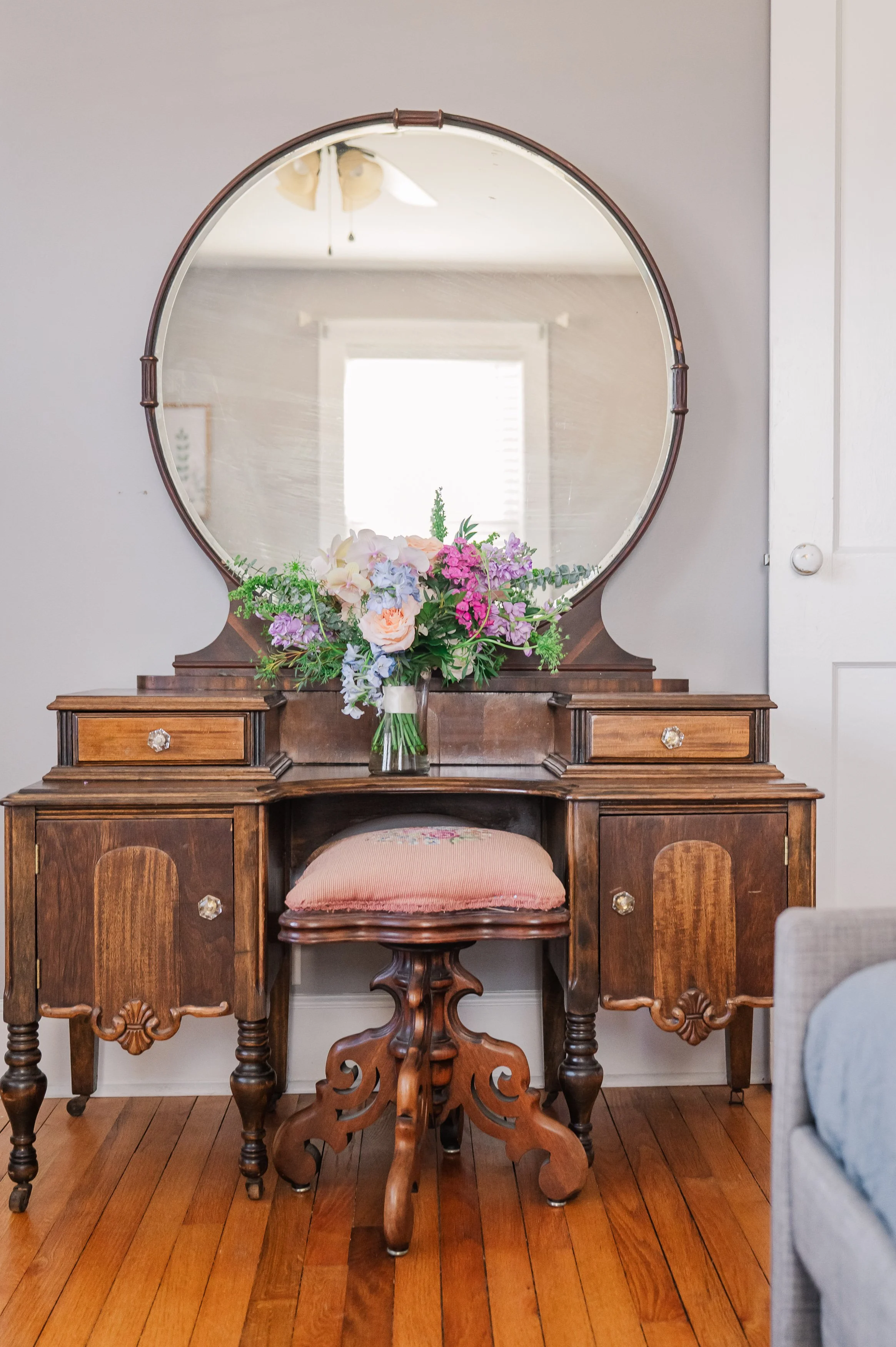 Vintage wooden dresser with a large round mirror, decorated with a bouquet of flowers on top, and matching vintage chair with pink cushion in front, all on hardwood floor.