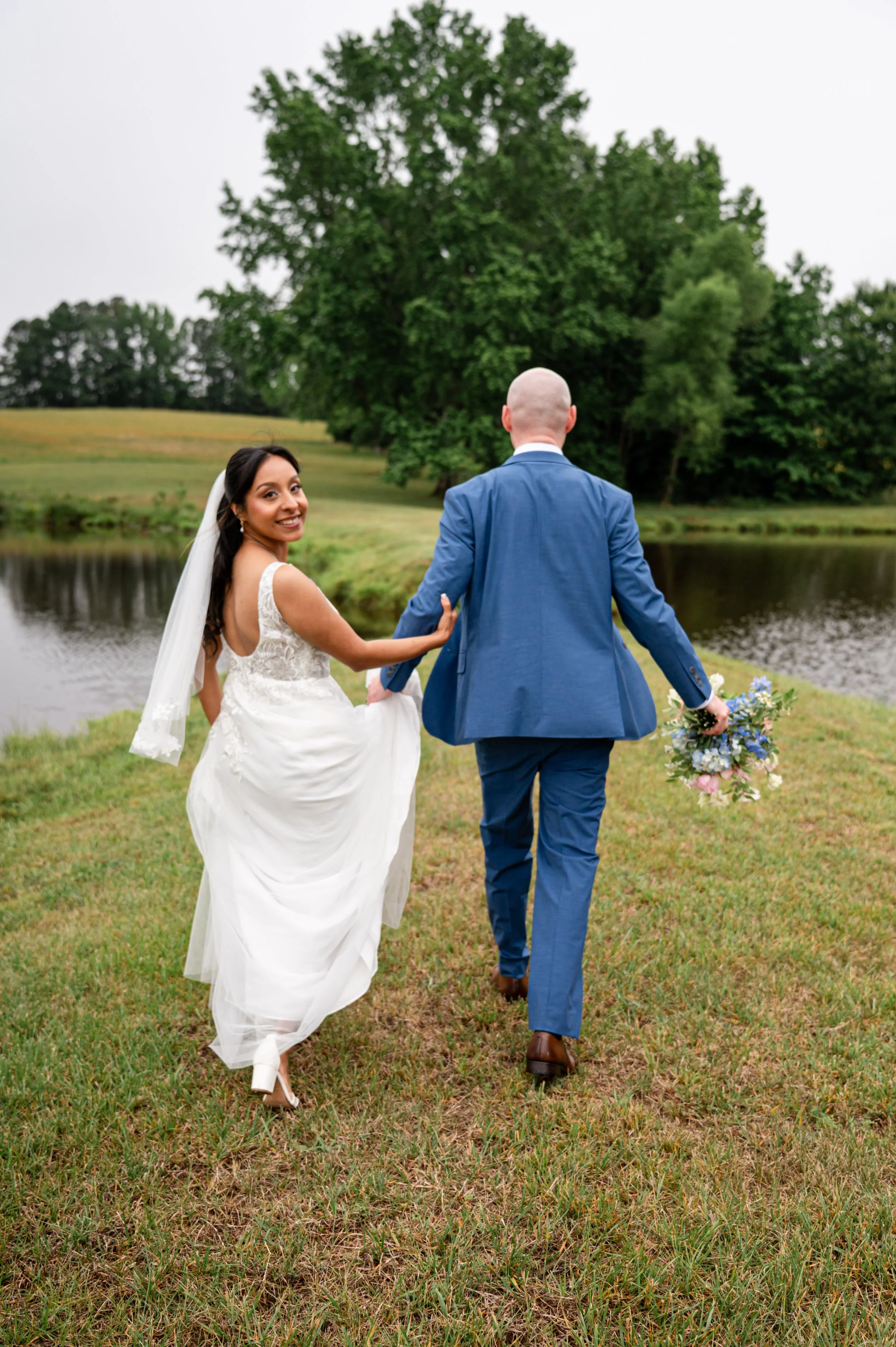 A happy bride in a white wedding dress and veil holding hands with a groom in a blue suit, walking by a pond with green grass and trees in the background on a cloudy day.