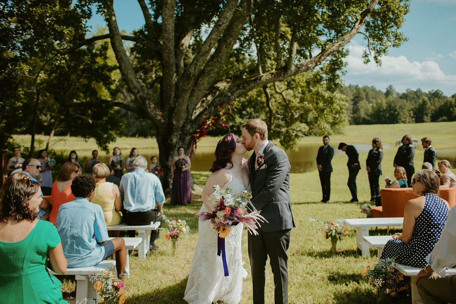 A wedding ceremony taking place outdoors under a large tree with a lake in the background. The bride and groom are kissing, with guests seated and standing around watching the scene. The bride is holding a colorful bouquet.