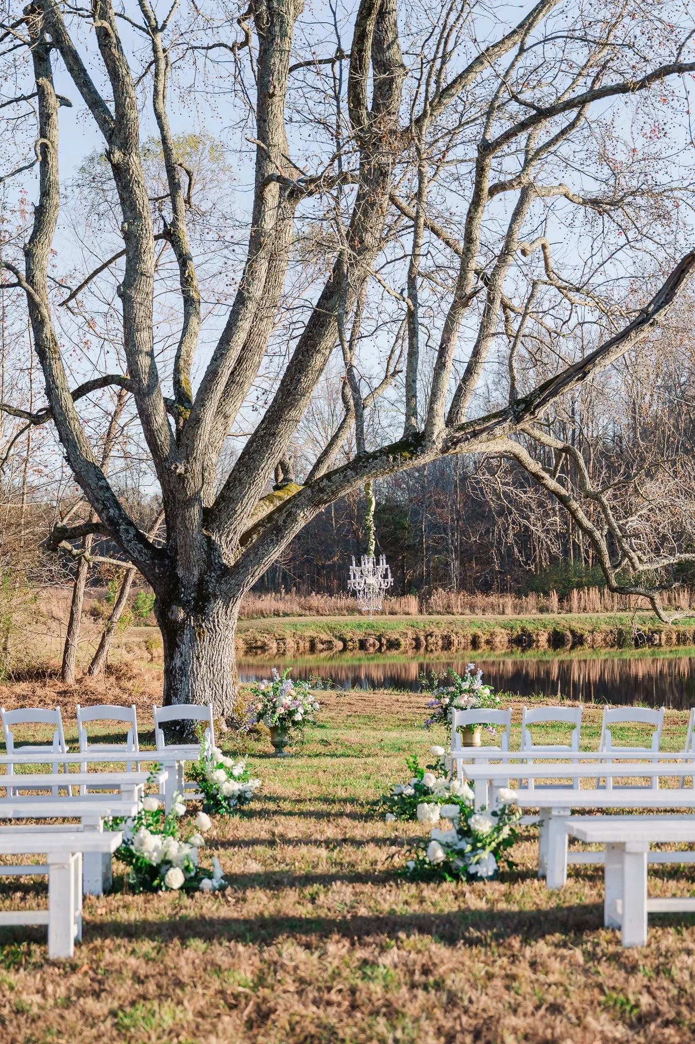 Outdoor wedding setup featuring white chairs and floral arrangements near a large tree with a lake in the background and a festive chandelier hanging from the tree branches at Sable and Vine, a wedding and event venue in Stem, North Carolina