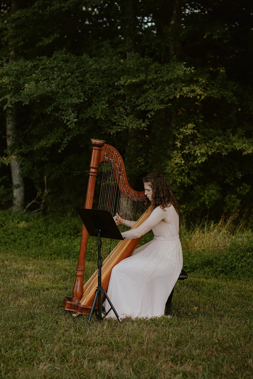 A woman in a long, light-colored dress playing a harp outdoors in a grassy area with trees in the background.