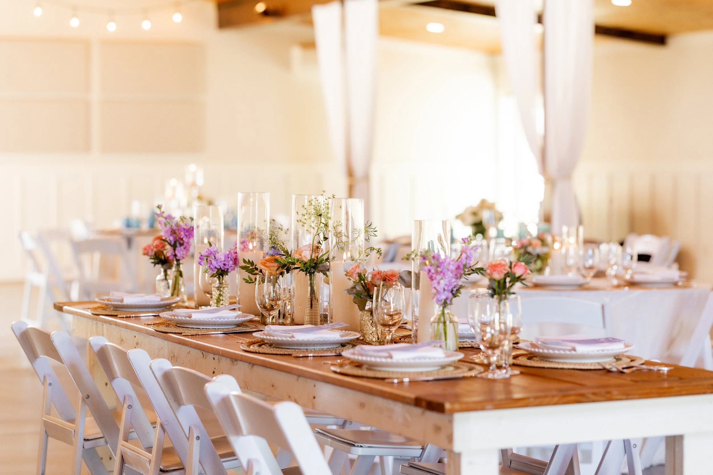 A decorated banquet table with floral centerpieces, candles, and place settings, set for a celebration in a bright, airy venue with exposed wooden beams.