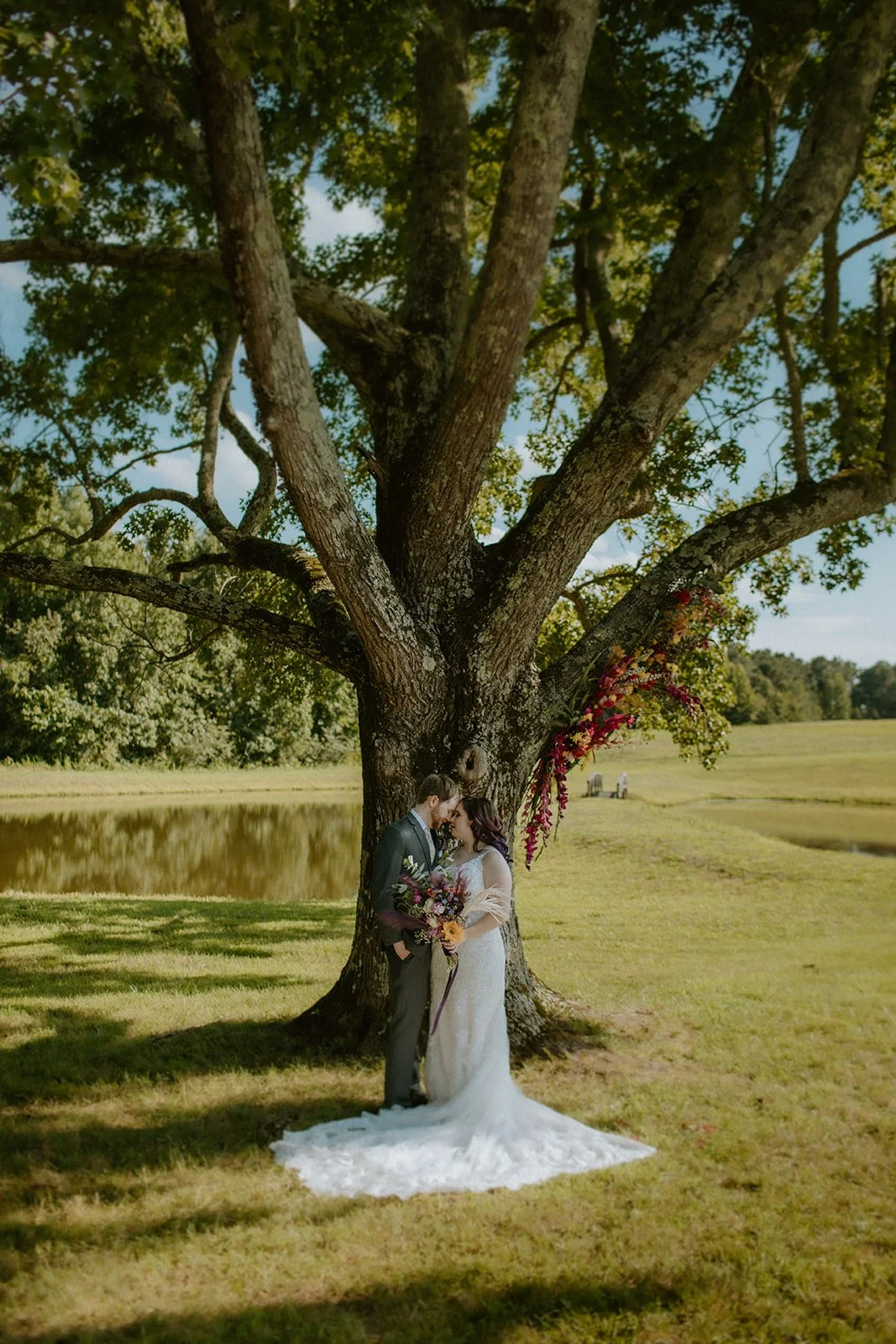 A bride and groom standing close together under a large tree, holding a bouquet of flowers, on the grass near a pond in a park-like setting.