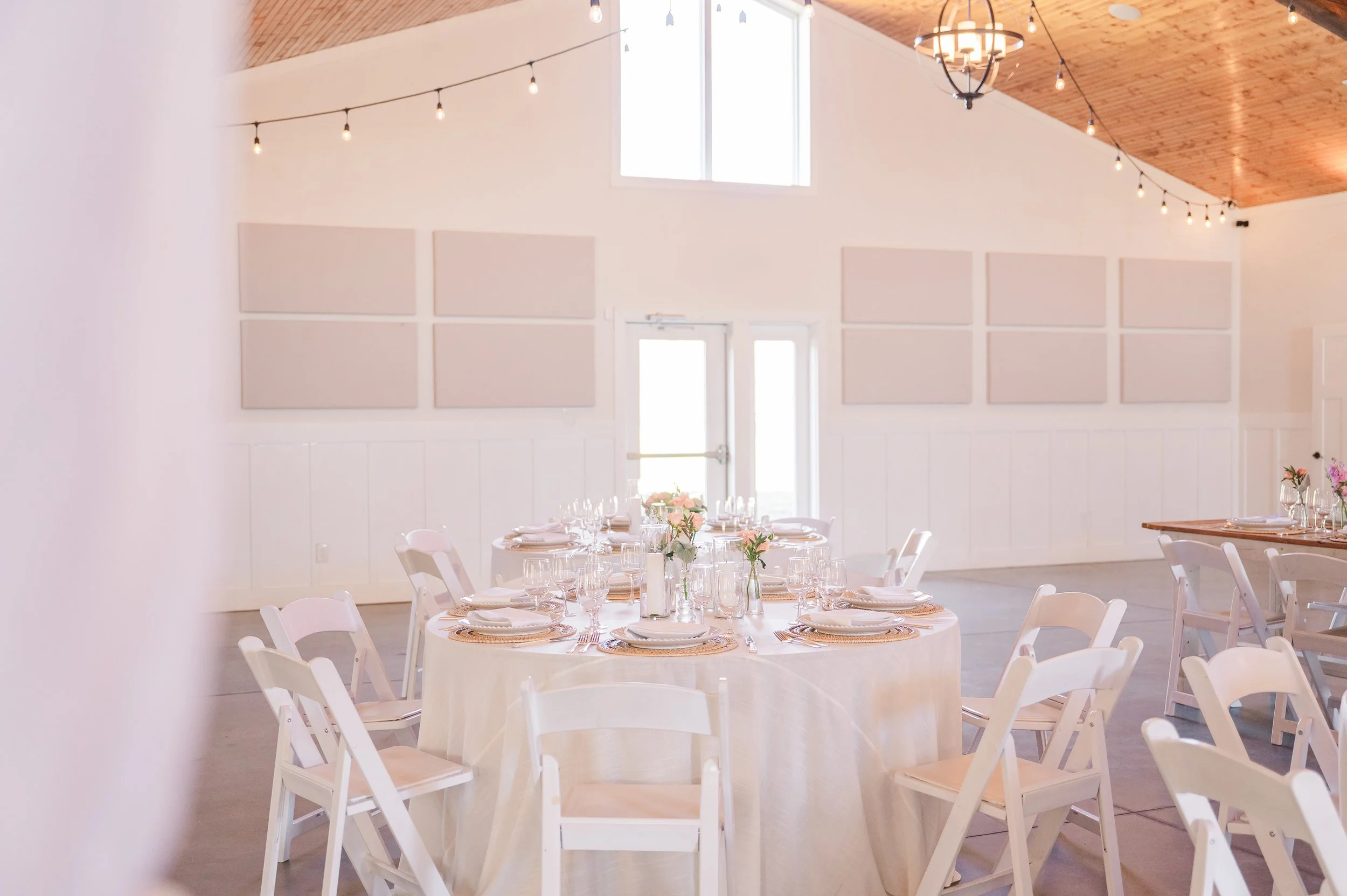 Wedding reception setup with round tables decorated with white tablecloths, floral centerpieces, and neatly arranged glassware and silverware in a bright, airy room with high ceilings, large windows, and string lights.