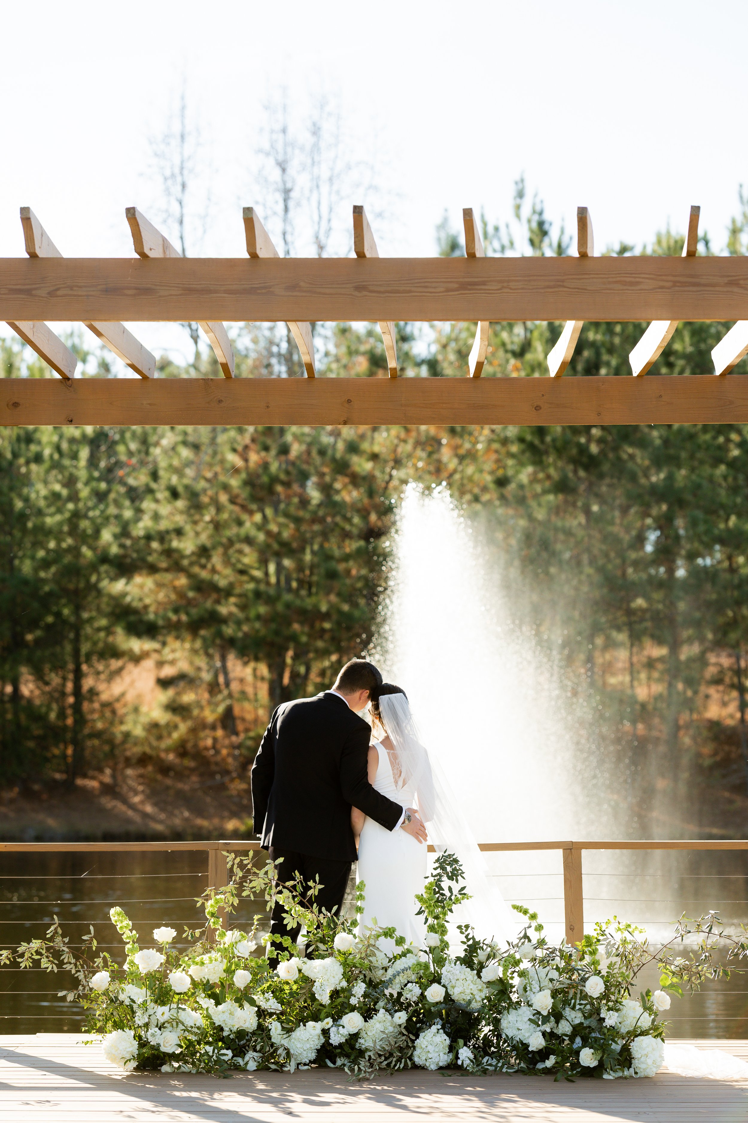 A bride and groom embrace during a wedding ceremony outdoors by a fountain, with a floral arrangement of white roses and greenery on a wooden platform, under a wooden pergola.
