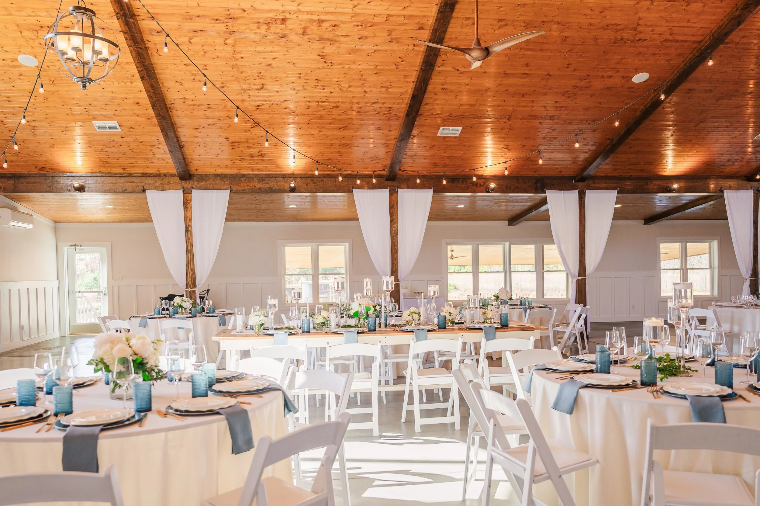 Elegant banquet hall with round tables set with white tablecloths, floral centerpieces, and place settings, featuring a wooden ceiling with string lights and white curtains.