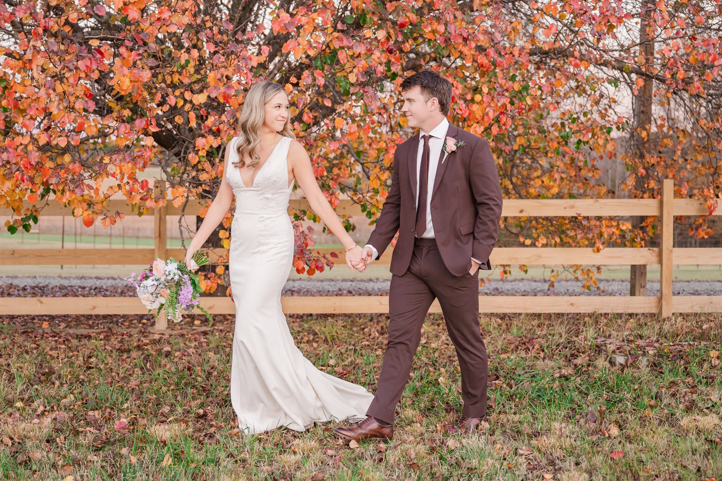 A bride and groom holding hands outdoors in front of a tree with red and orange autumn leaves, during a wedding.