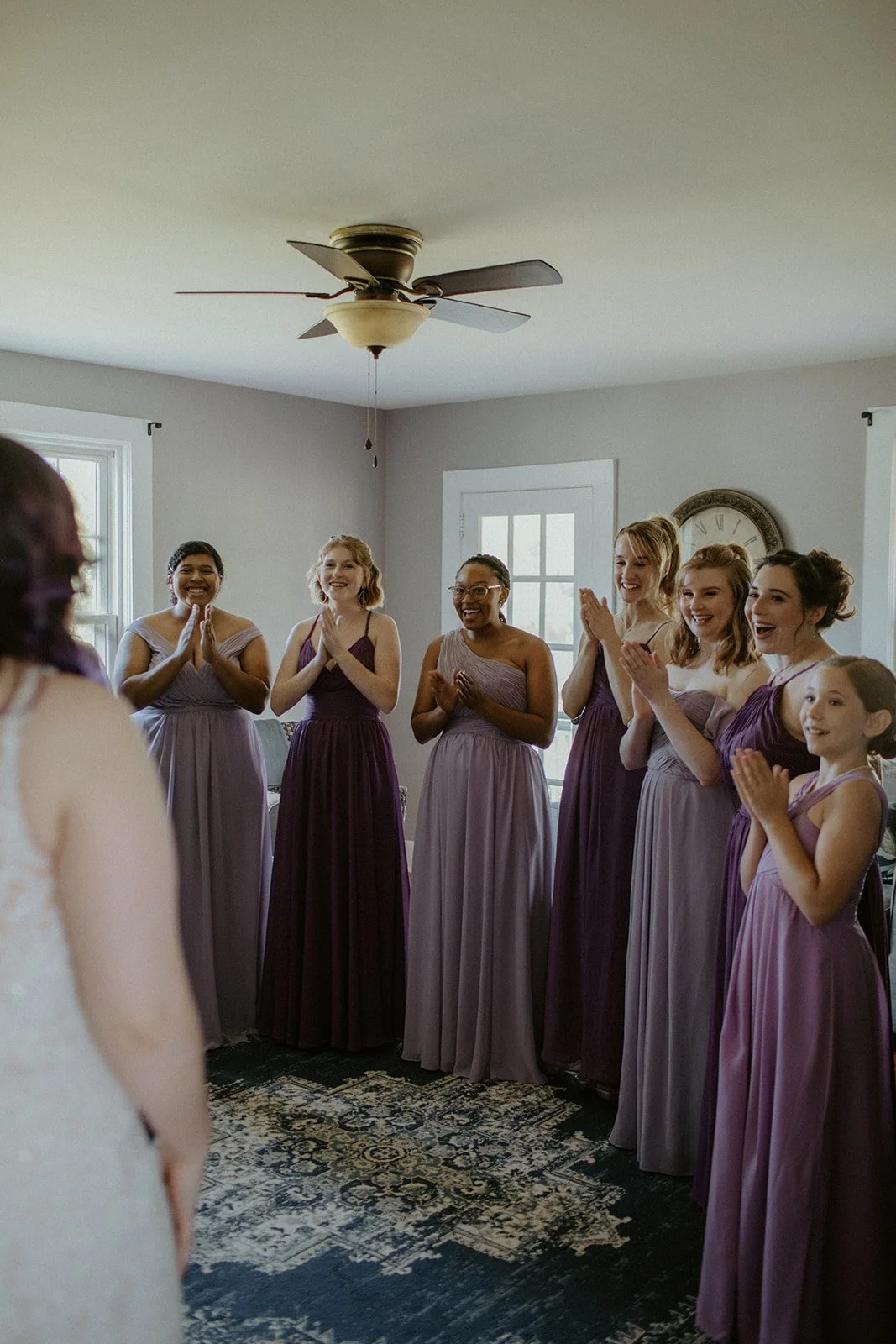 Group of women and a young girl in purple dresses smiling and clapping in a well-lit room with a ceiling fan and windows.