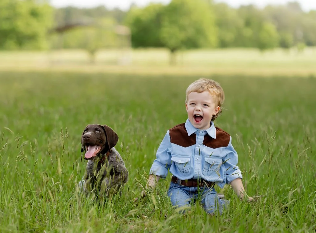 A young boy and a puppy sitting in a grassy field, smiling and enjoying outdoors.