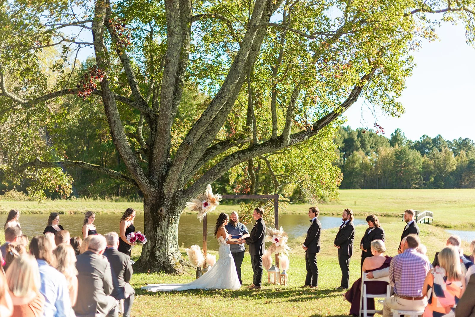 A wedding ceremony taking place outdoors under the Sable and Vine ceremony tree near the on-site pond with the bride and groom exchanging vows, officiant, wedding party and their family and friends
