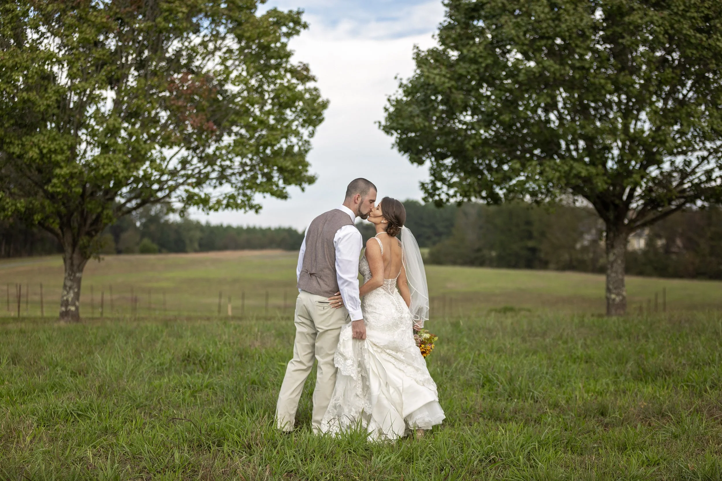 A bride and groom share a kiss outdoors under large trees, with green grass and an open field in the background during daytime.