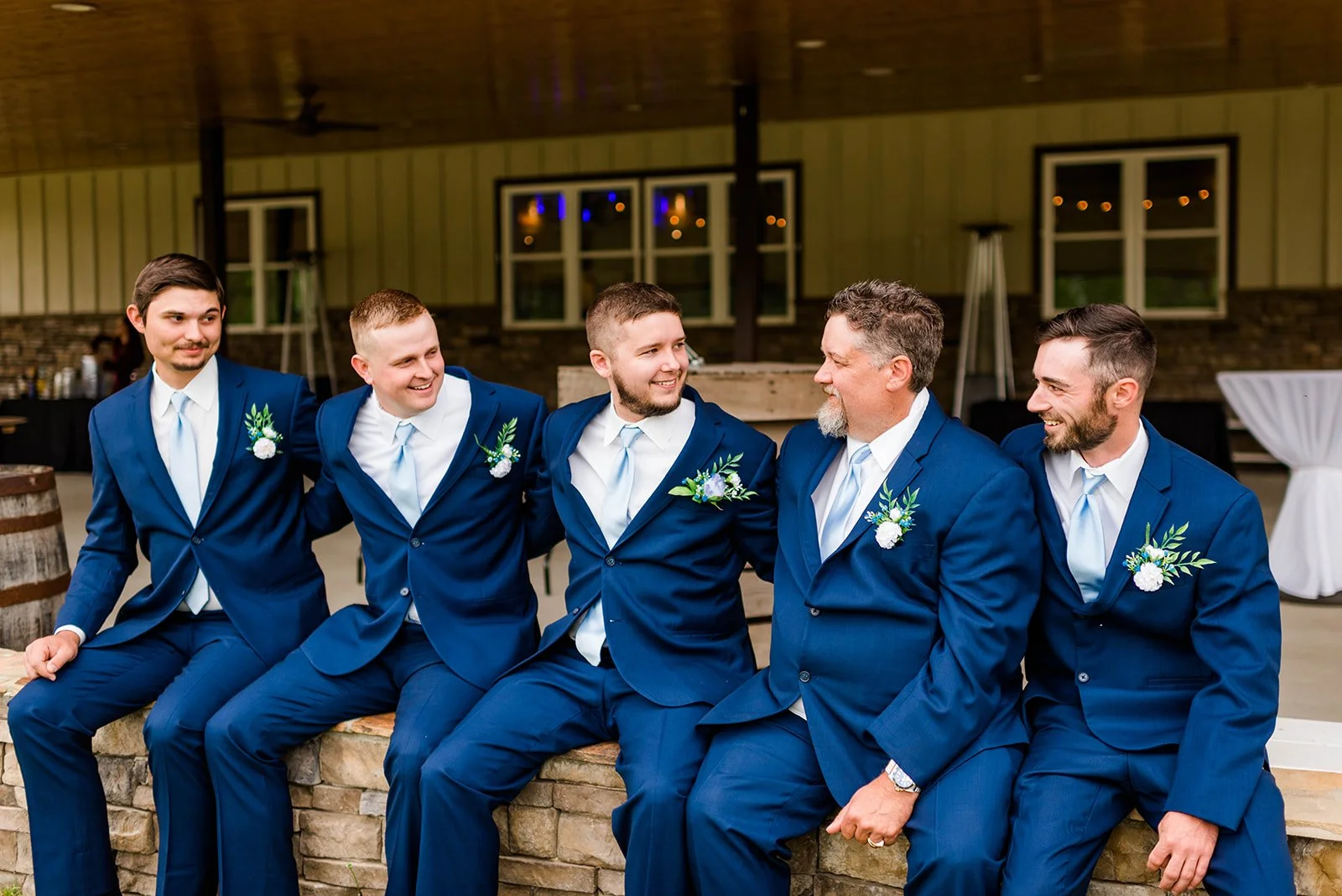 A group of five men in matching blue suits with boutonnières, sitting and smiling together at a wedding or formal event inside a rustic venue with wood-paneled walls and windows.