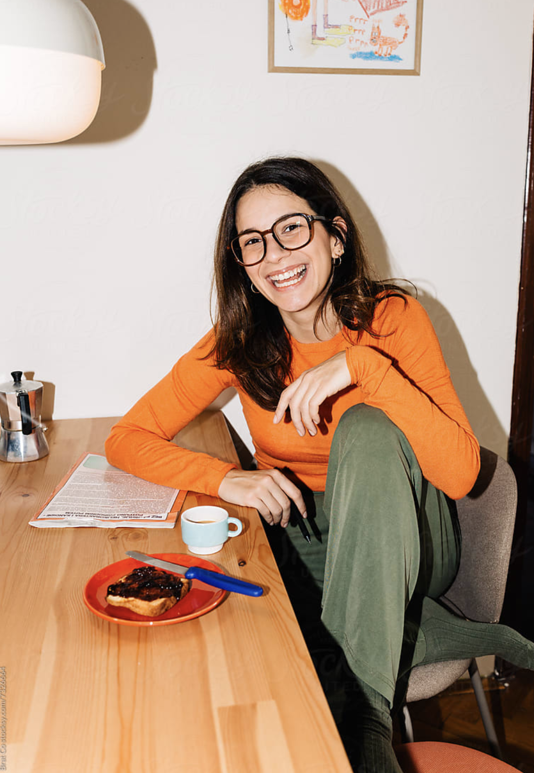 A woman with long dark hair, glasses, and hoop earrings, wearing an orange long sleeve shirt and green pants, sitting at a wooden table with a plate of toast with jam, a cup of coffee, a newspaper, and smiling at the camera in a cozy room with artwork on the wall.