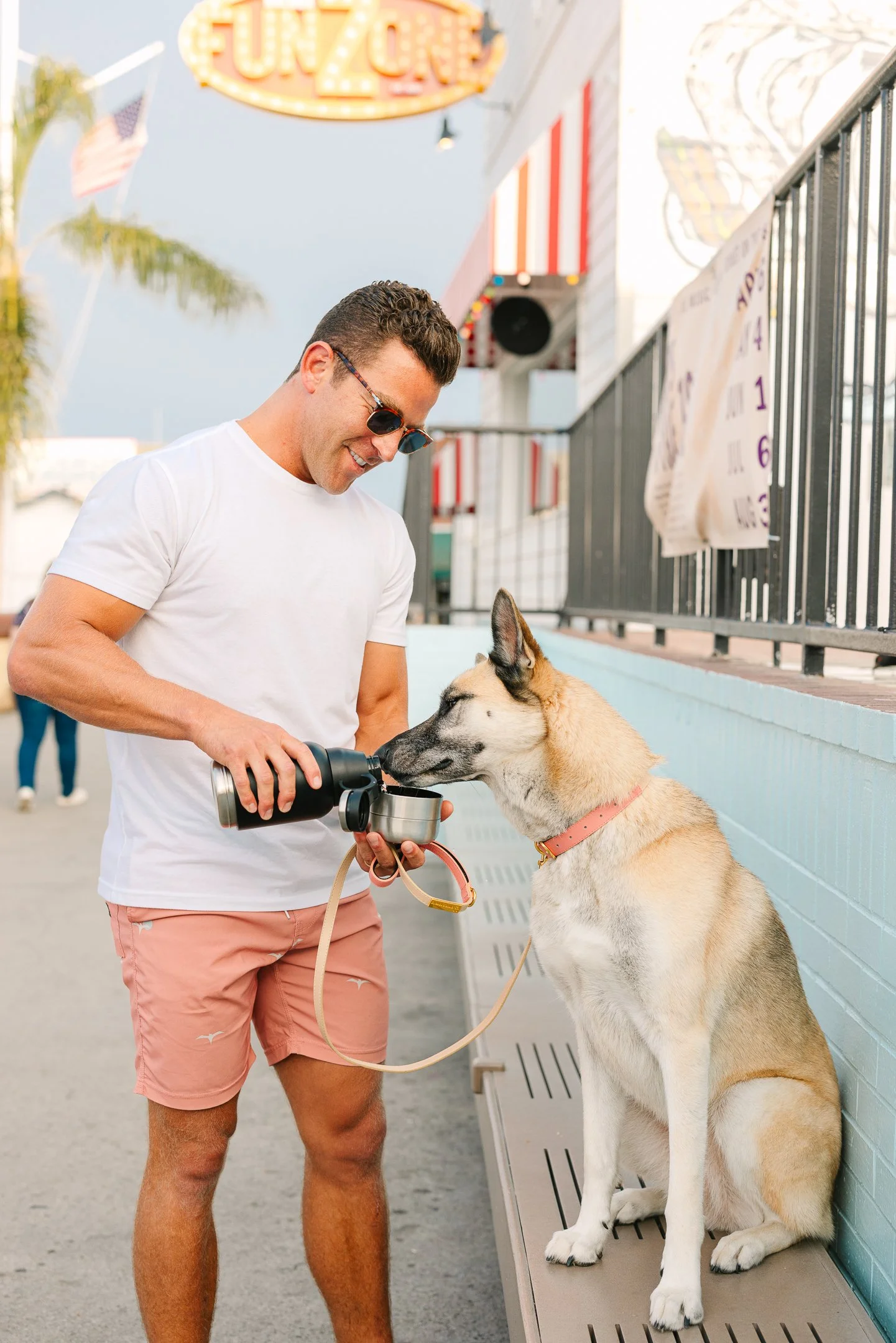 A man wearing sunglasses, a white t-shirt, and pink shorts is feeding a large dog with a food bowl outside at a boardwalk or amusement park. The dog is sitting on a bench, and there is a sign that says "Coney" in the background.