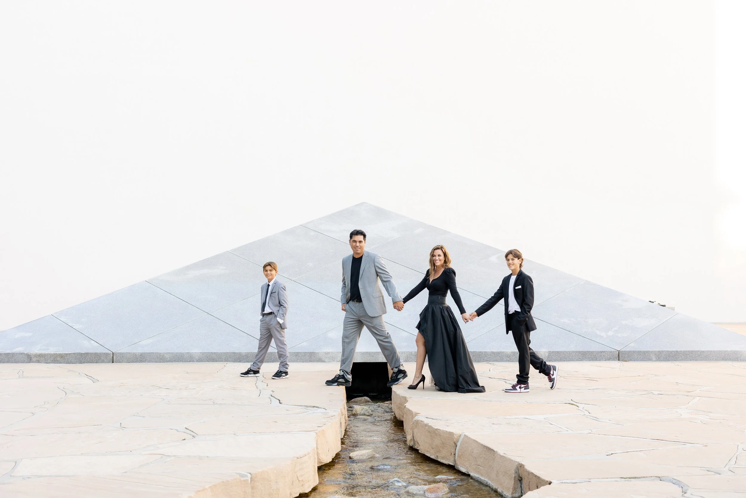 A group of five diverse people, dressed in formal and semi-formal attire, holding hands and walking across a stone bridge, with a modern, minimalistic building in the background.