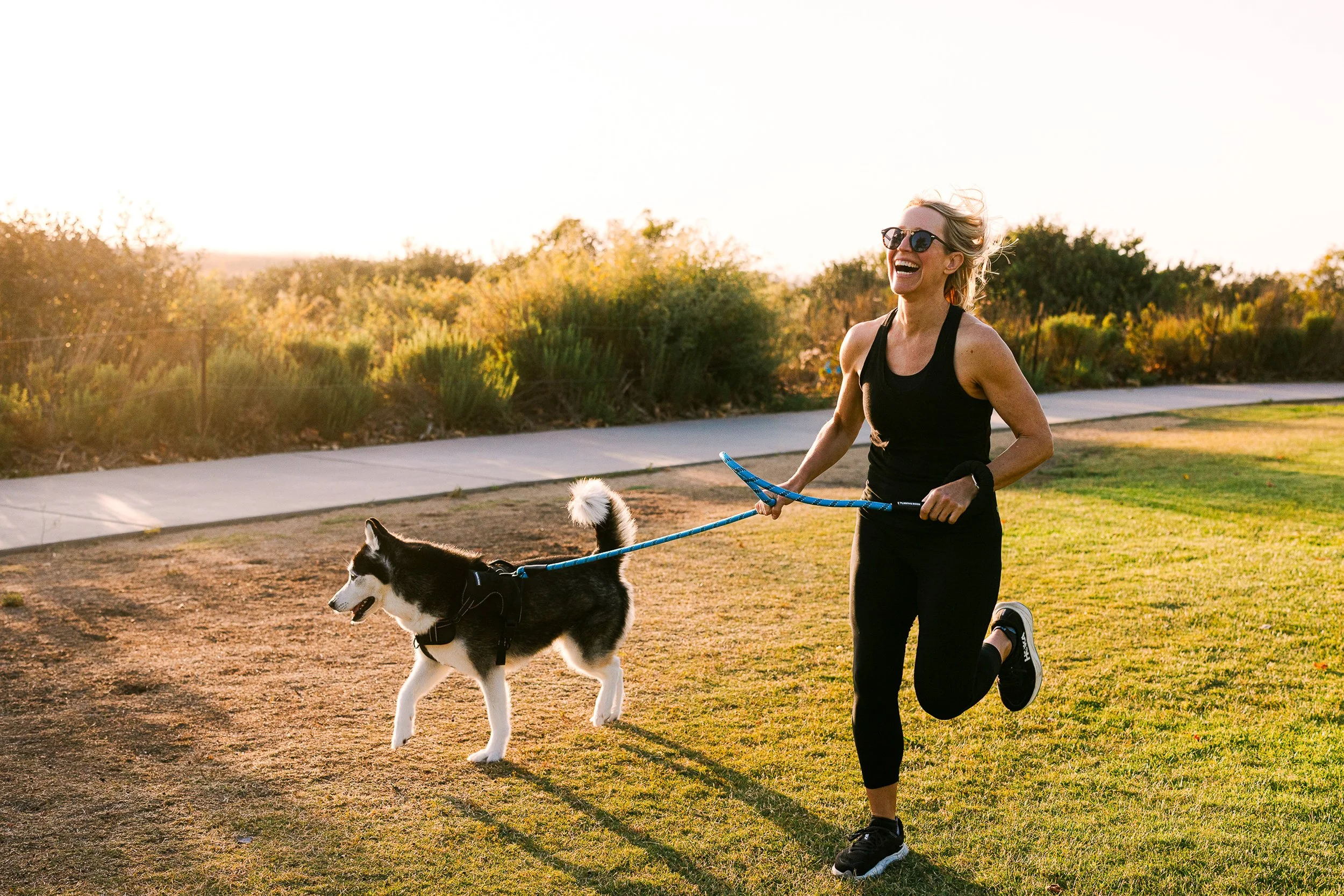 A woman running outdoors with her Siberian Husky dog during sunset, wearing black athletic clothes and sunglasses, on a grassy path with trees in the background.