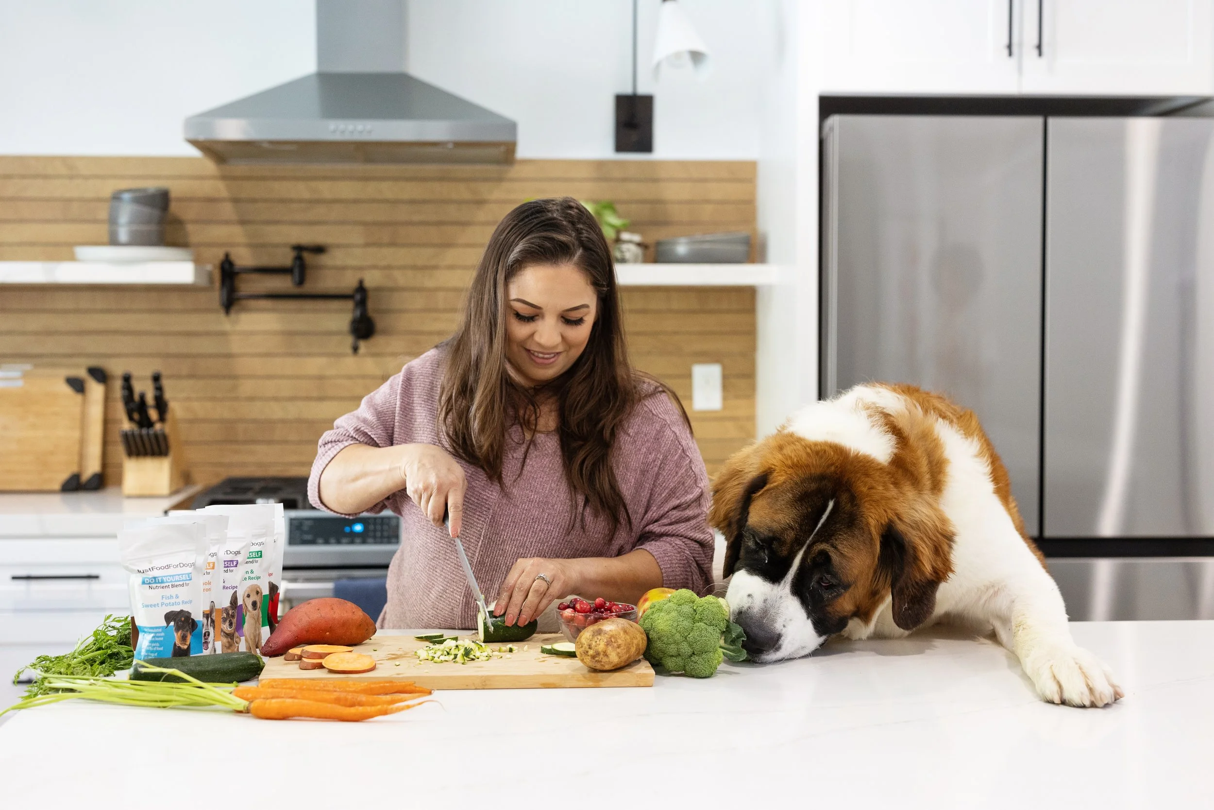 A woman and a large Saint Bernard dog preparing vegetables in a modern kitchen. There are carrots, sweet potatoes, broccoli, cucumbers, and strawberries on the counter, along with pet food bags.
