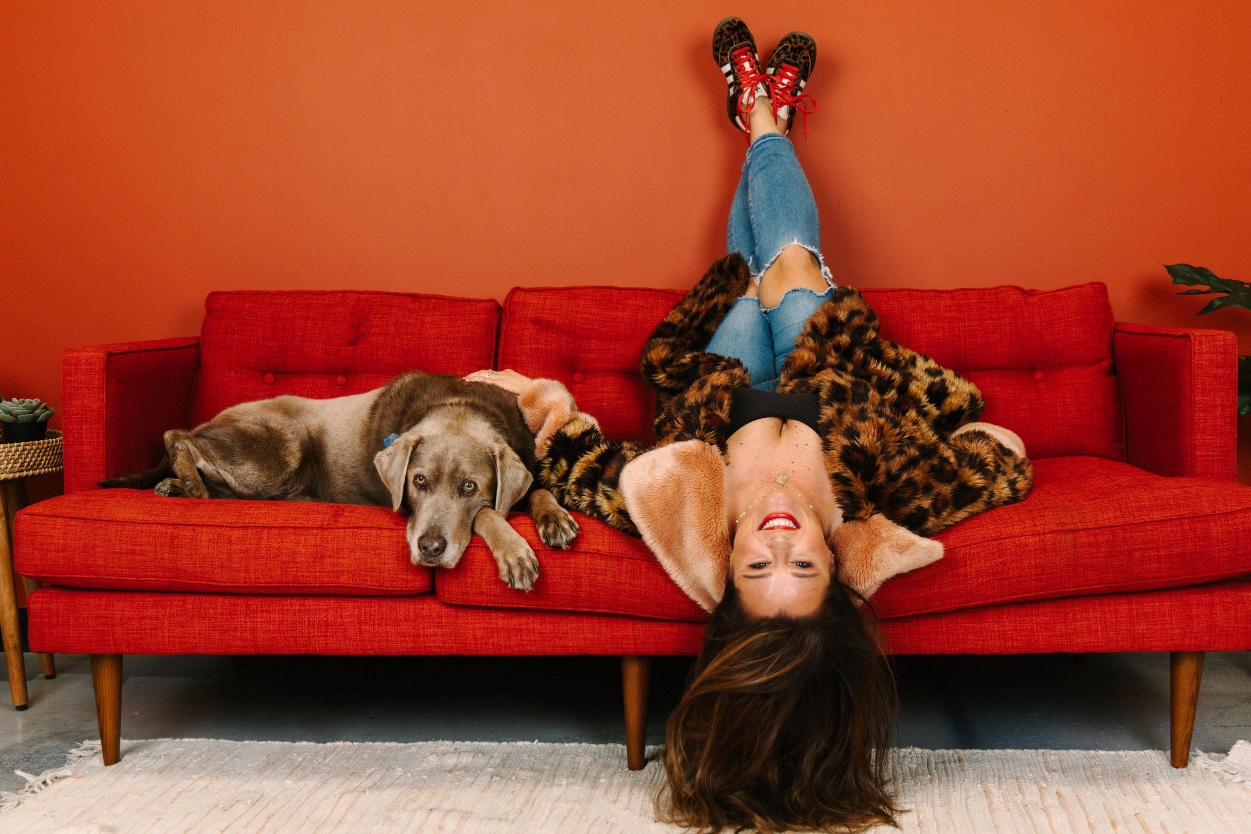 Woman lying upside down on a red sofa, with her head on the floor and her legs raised against the wall. She is smiling, wearing a leopard print coat, ripped jeans, and red shoes. There are two dogs resting on the sofa beside her, one as a Husky mix and the other as a Labrador mix. The room has a teal plant and a small succulent on a side table.