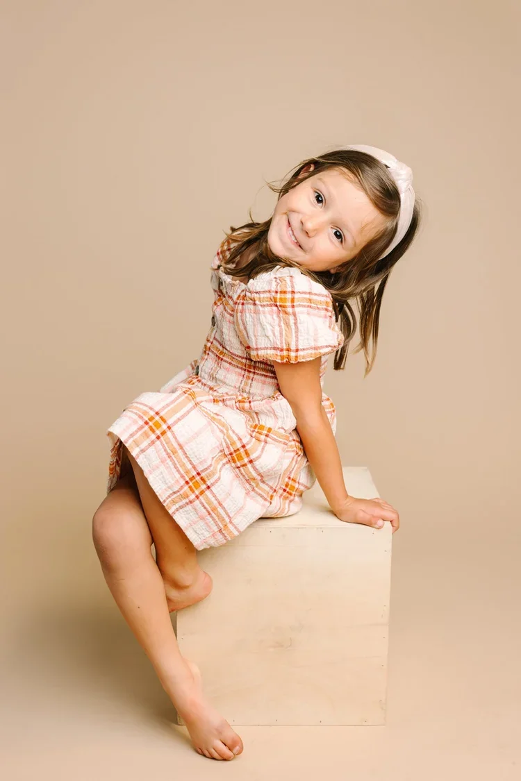 Young girl with brown hair wearing a pink and orange plaid dress, sitting on a wooden block, smiling and tilting her head to the side, against a beige background.