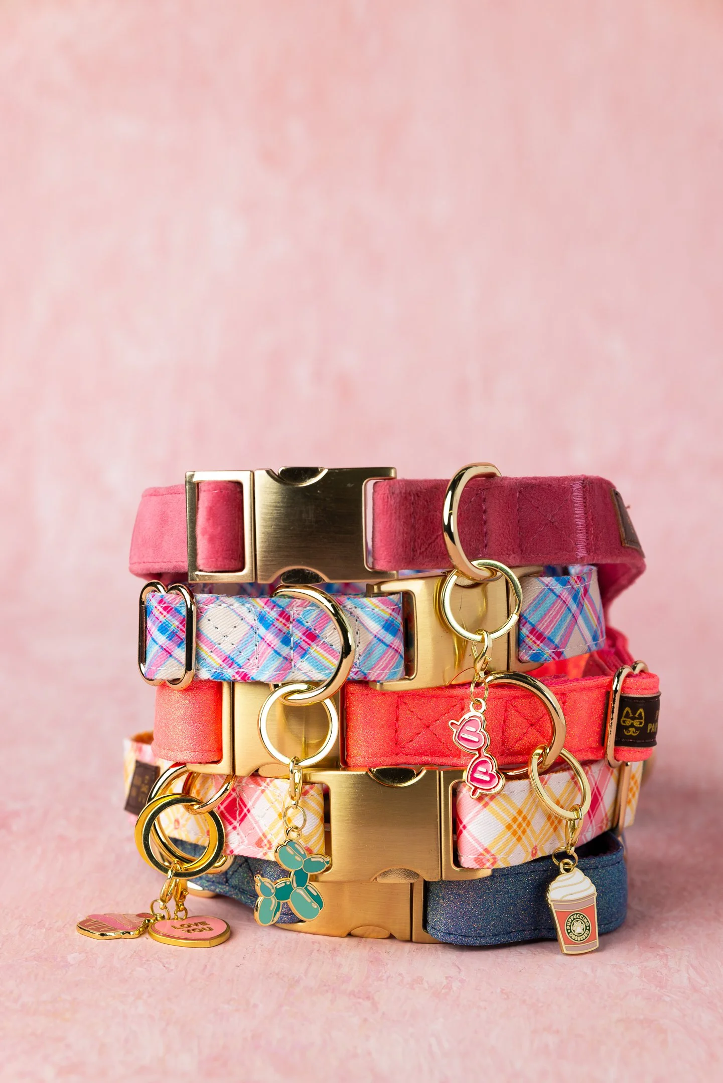 Stacked colorful dog collars with various charms on a gold stand against a pink background.