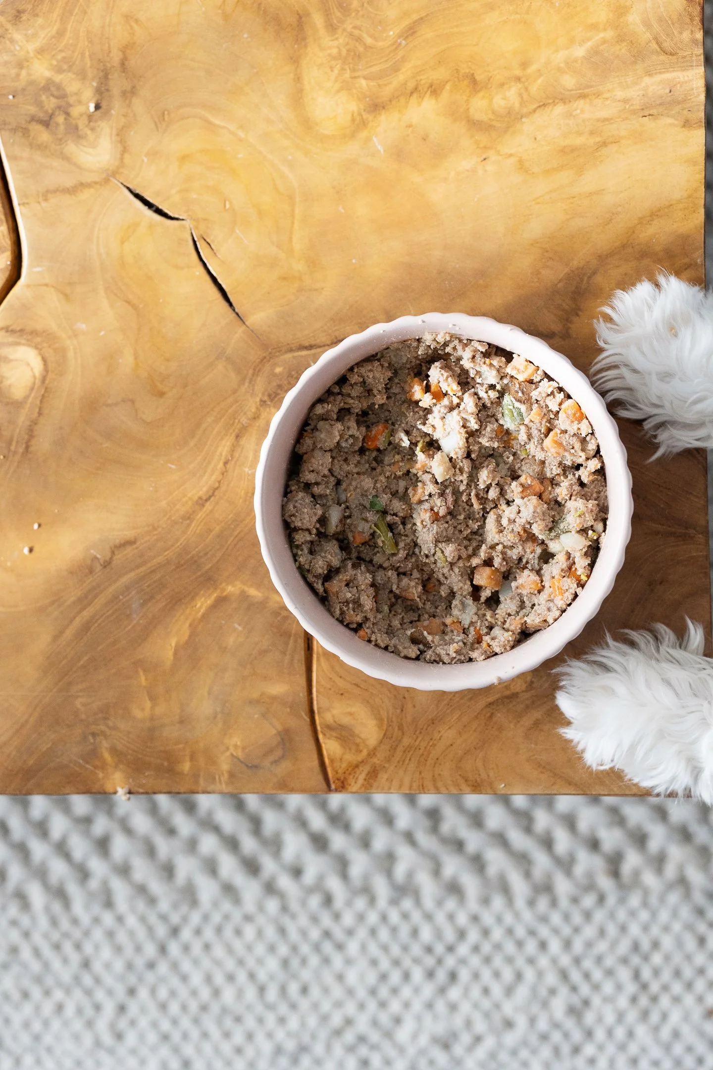 A bowl of ground meat and vegetable mixture on a wooden table, with a small dog partially visible on the right side.