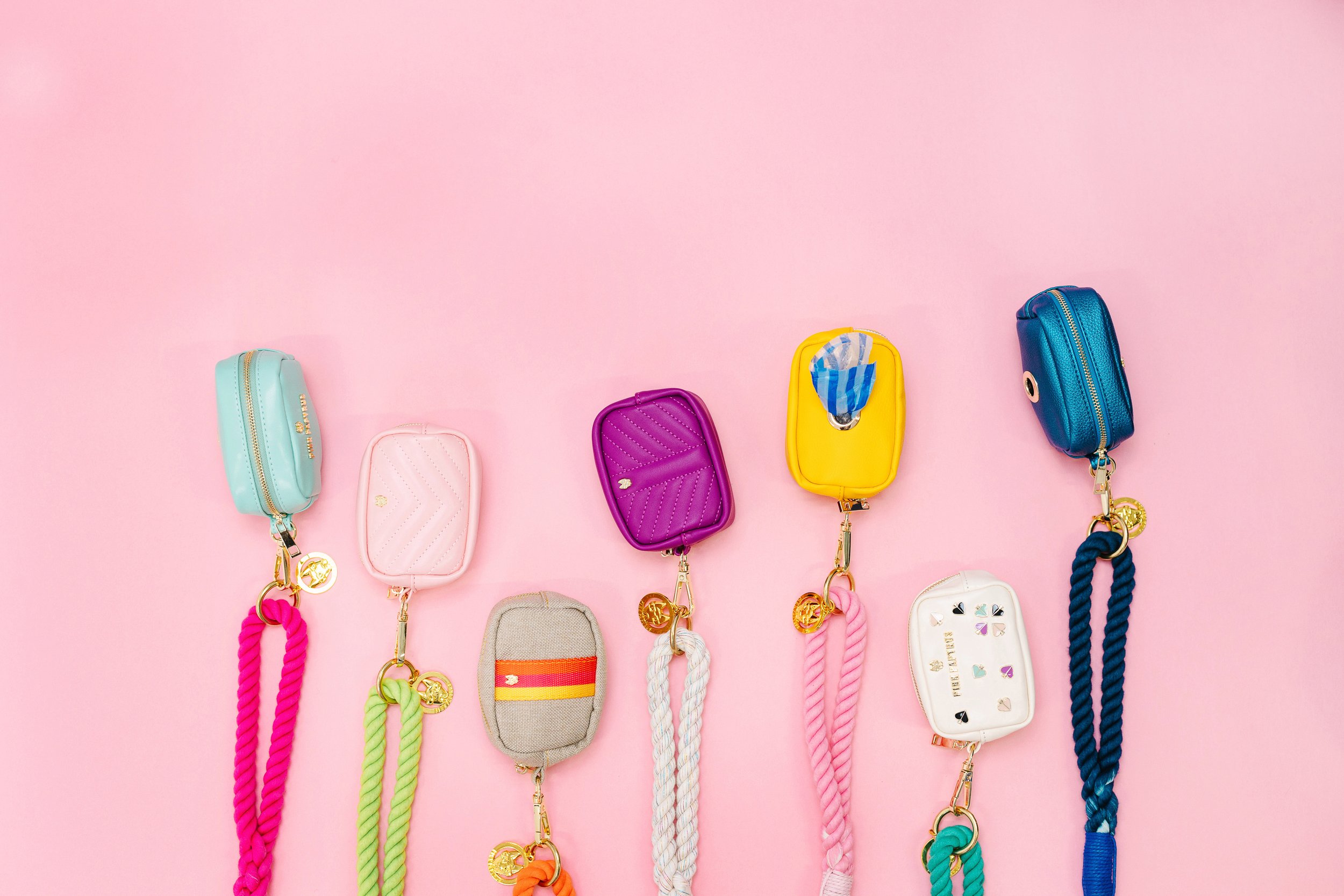 Colorful miniature handbags with matching braided straps and gold charms, arranged in a row against a pink background.
