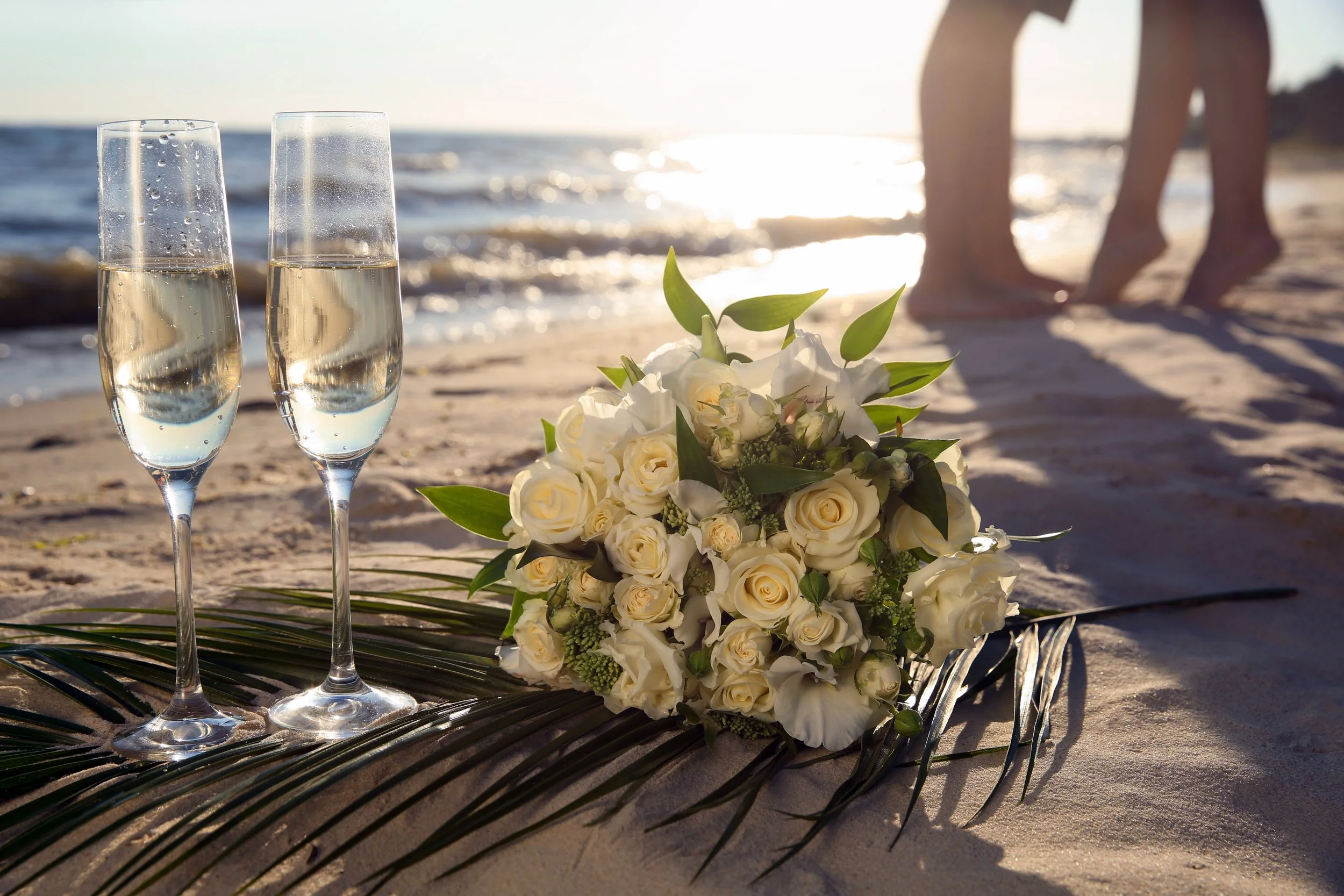 A bouquet of white roses and flowers lying on the sand with two glasses of champagne, at a beach during sunset.