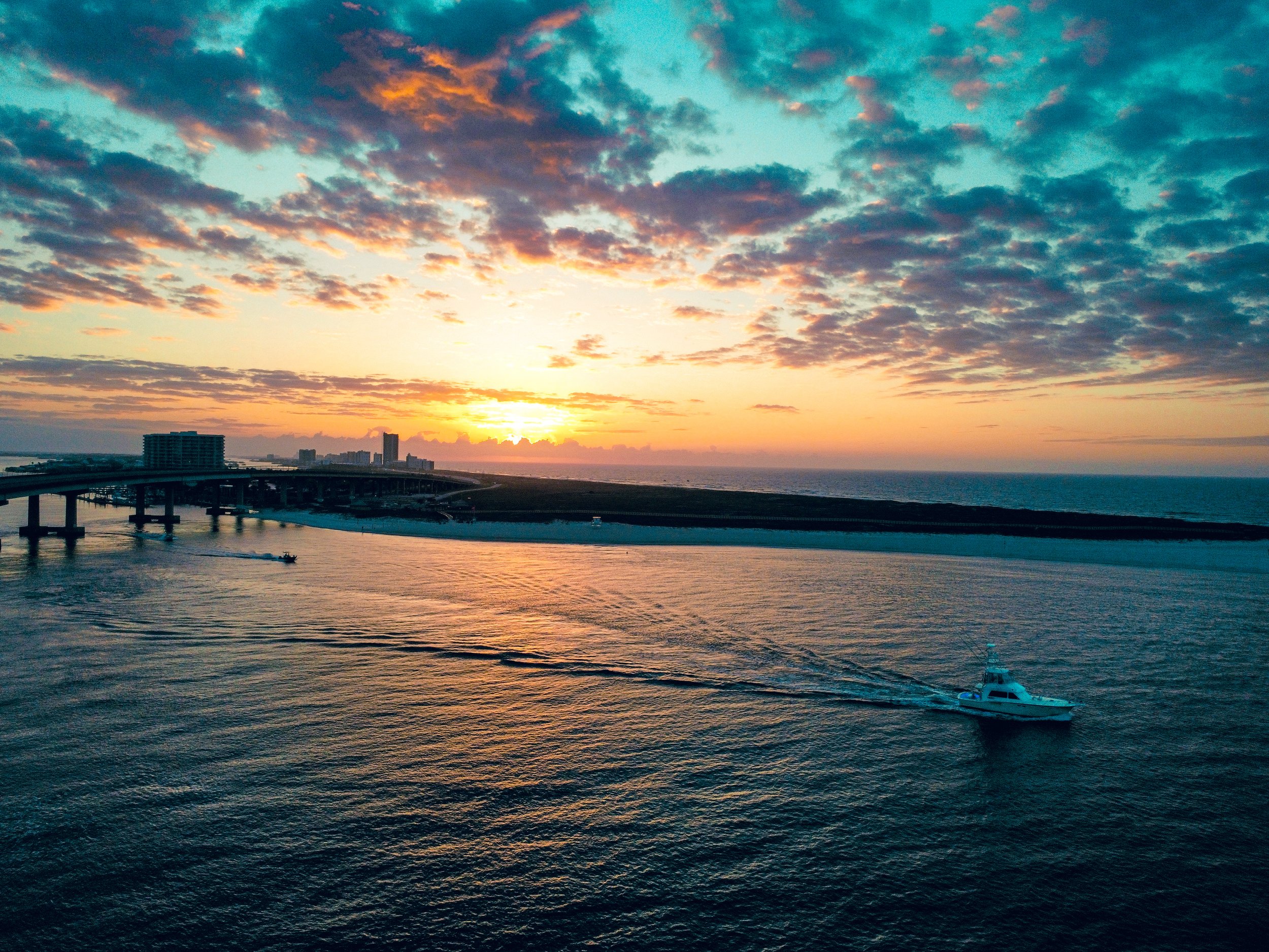 Sunset over the ocean with a boat sailing, a bridge, and buildings in the distance under a partly cloudy sky.