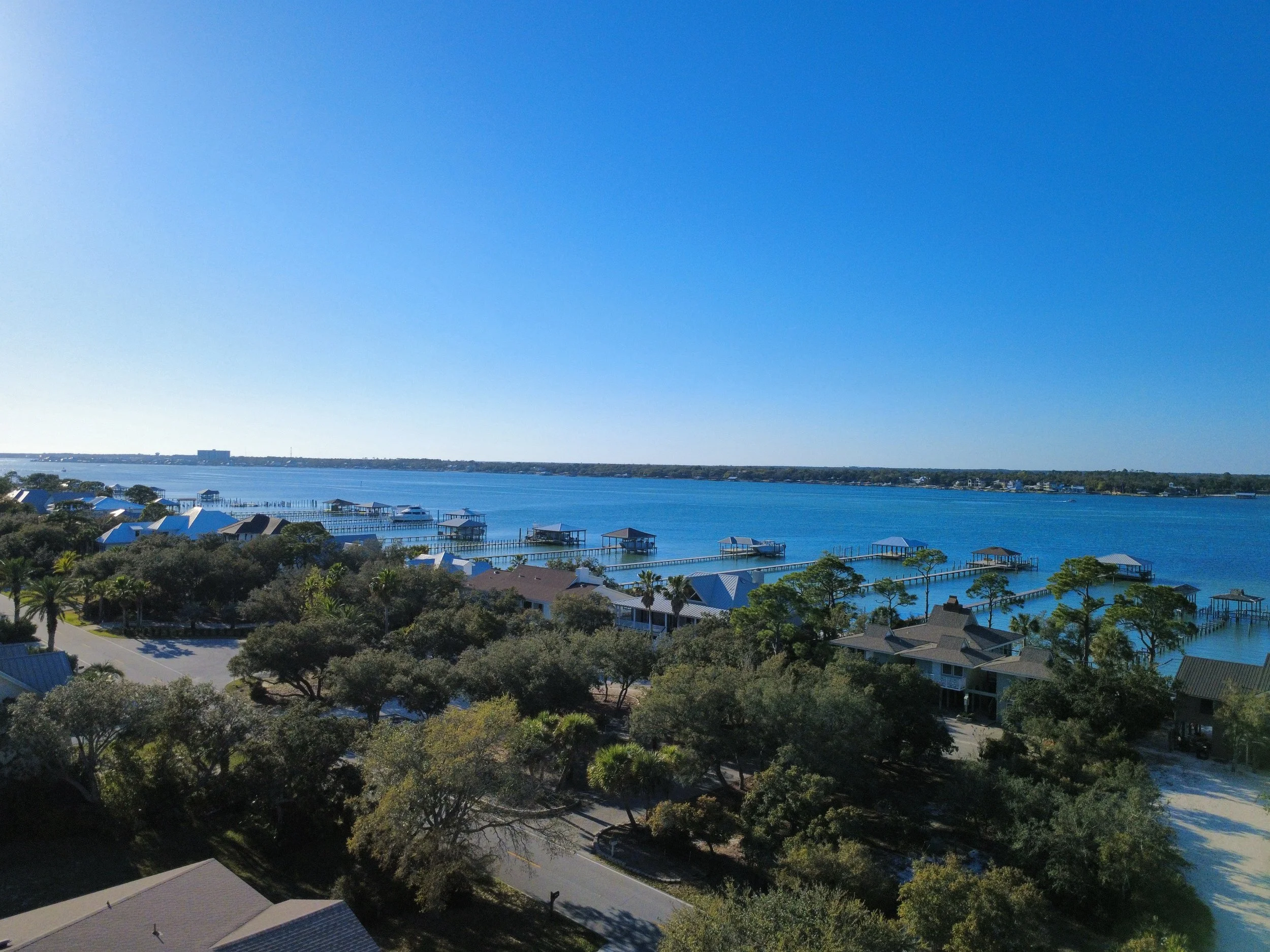 Aerial view of a waterfront area with trees, houses, and boats at dock, under a clear blue sky.