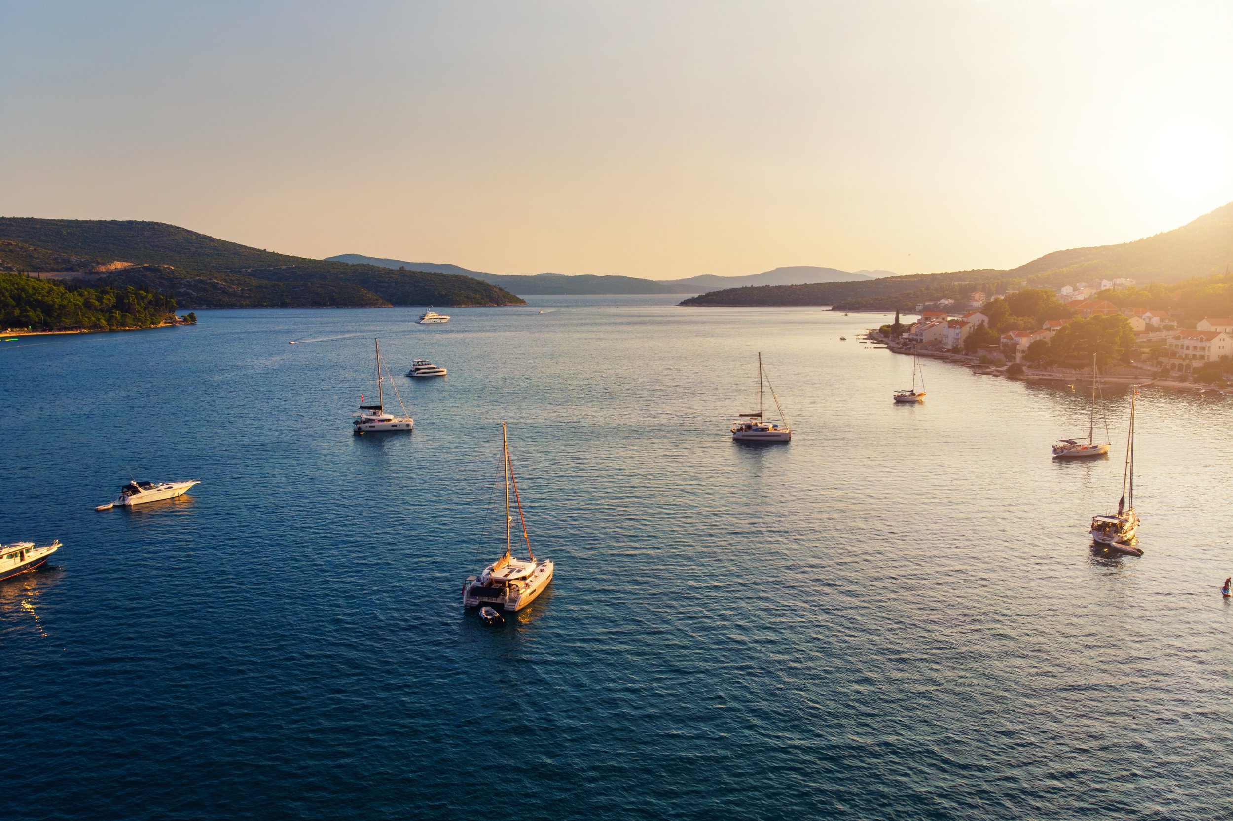 Boats floating on calm water near a coastal town during sunset or sunrise, with hills in the background.