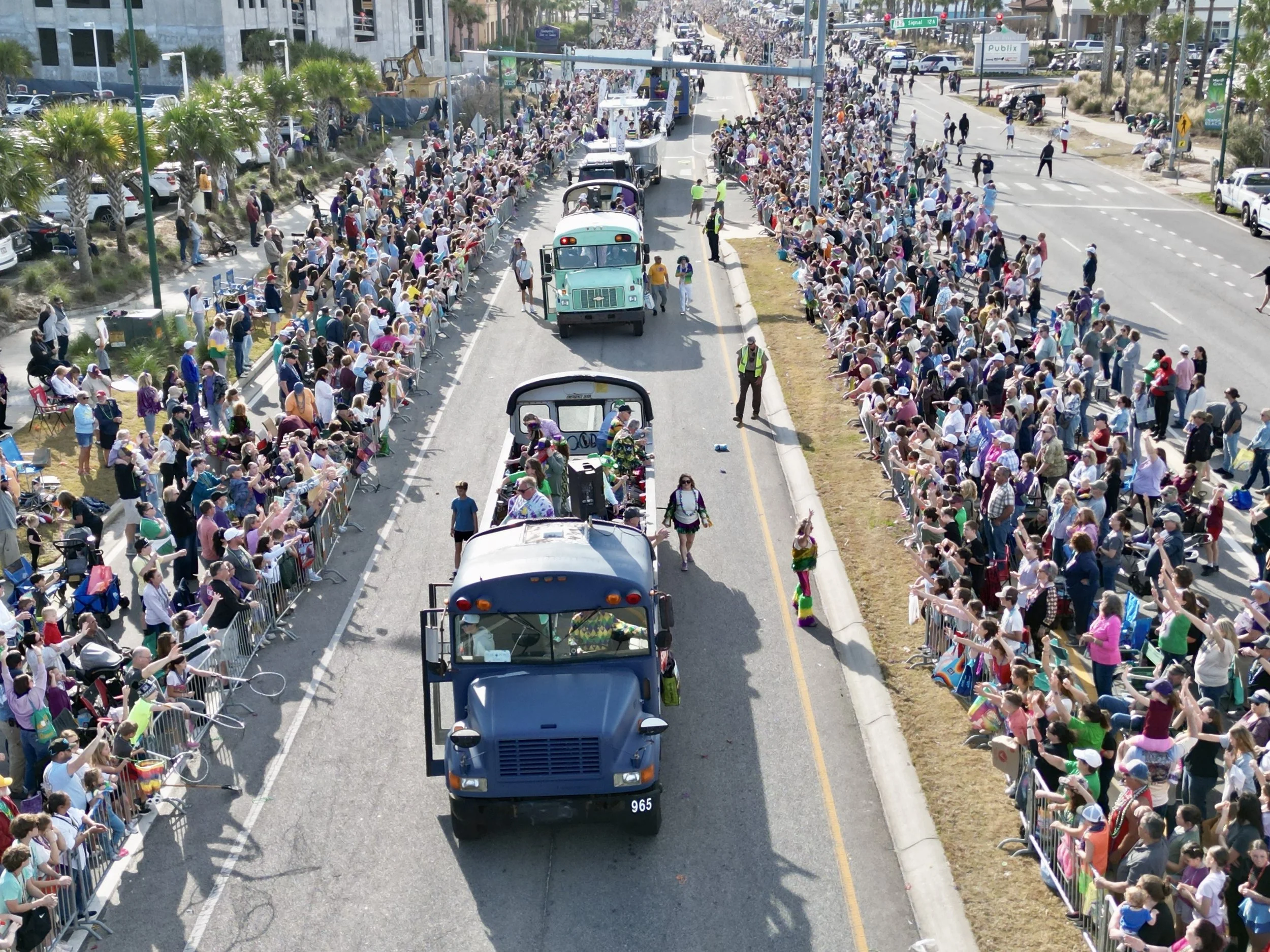 Crowd of people lined up on both sides of the street, participating in a parade with various decorated vehicles and festivities on a sunny day.