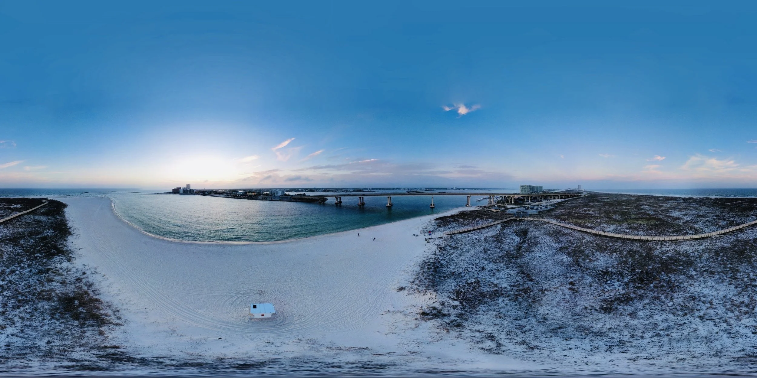 A panoramic view of a coastal area with a sandy beach, a pier extending into calm water, a small white hut on the beach, and snow-covered dunes and vegetation. The sky is clear with a few scattered clouds and the sun is setting or rising on the horiz