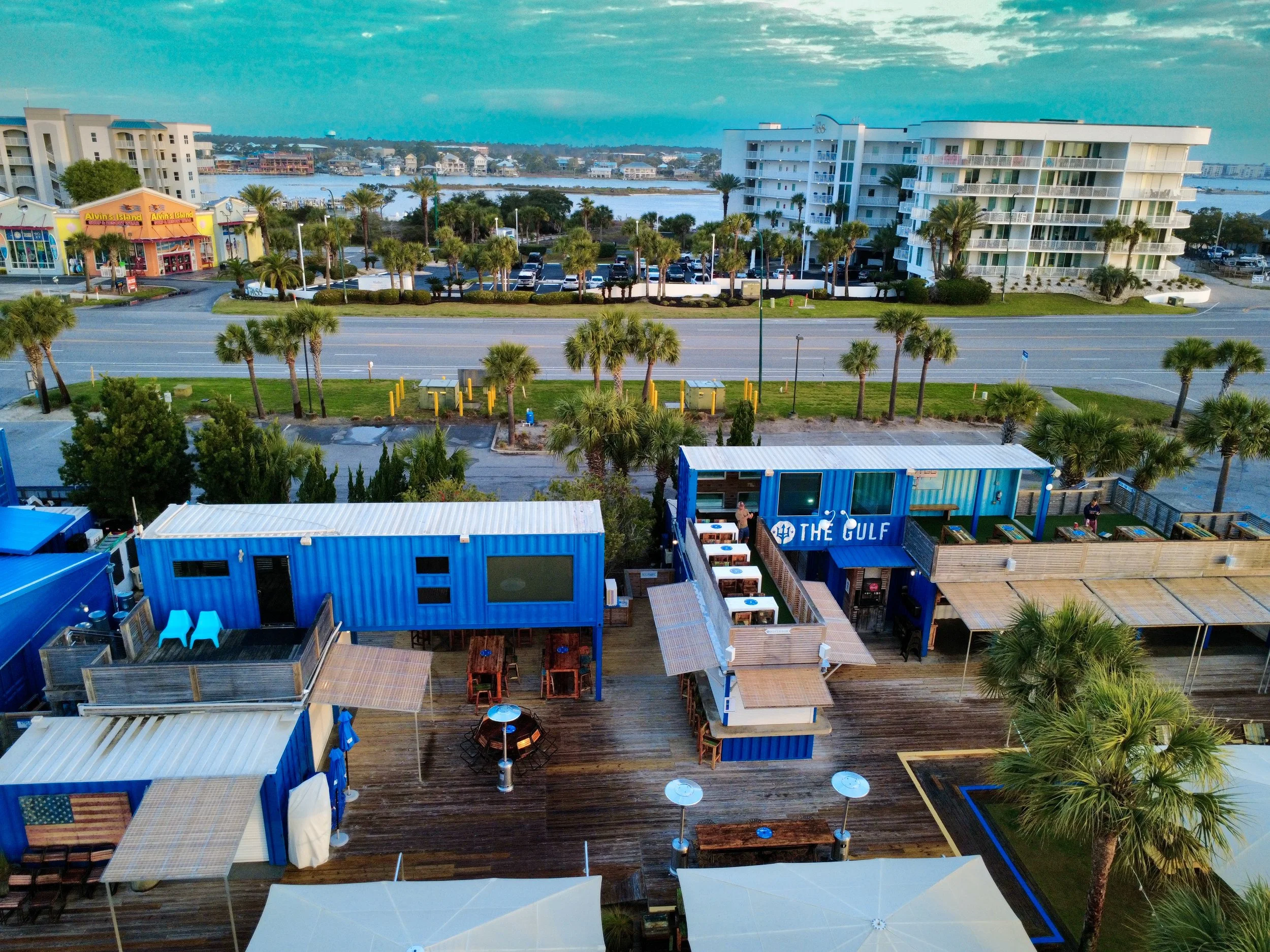 Aerial view of a tropical waterfront area with restaurants made from shipping containers, palm trees, a road, and water in the background.