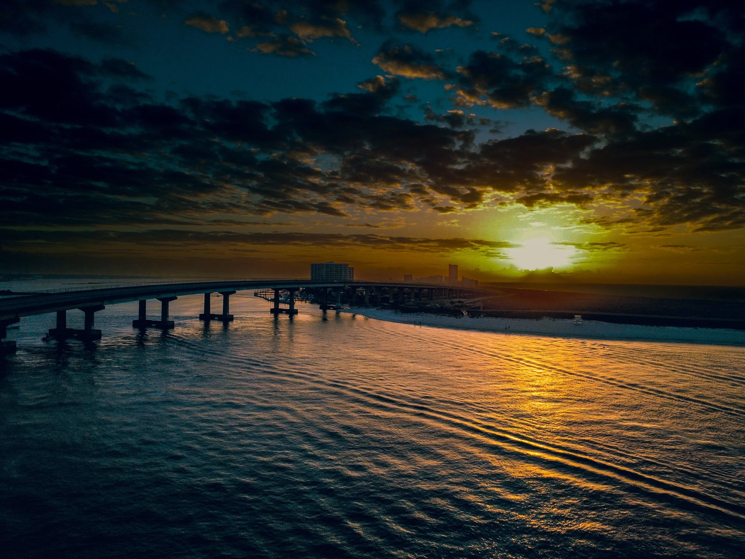 Sunset over a bridge crossing a body of water, with the sky partly cloudy and city buildings silhouetted in the distance.