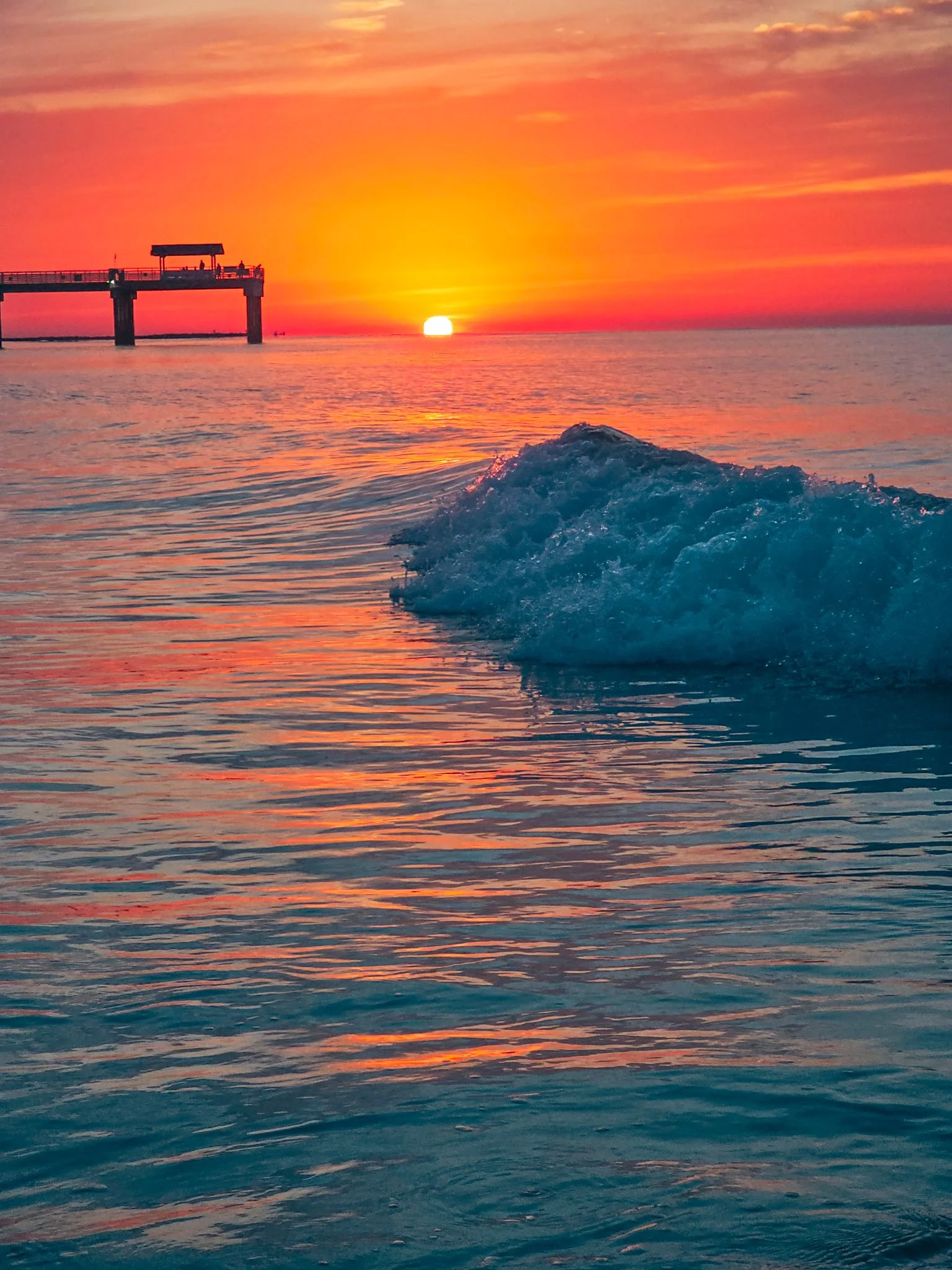 Sunset over the ocean with a pier in the distance and gentle waves in the foreground.