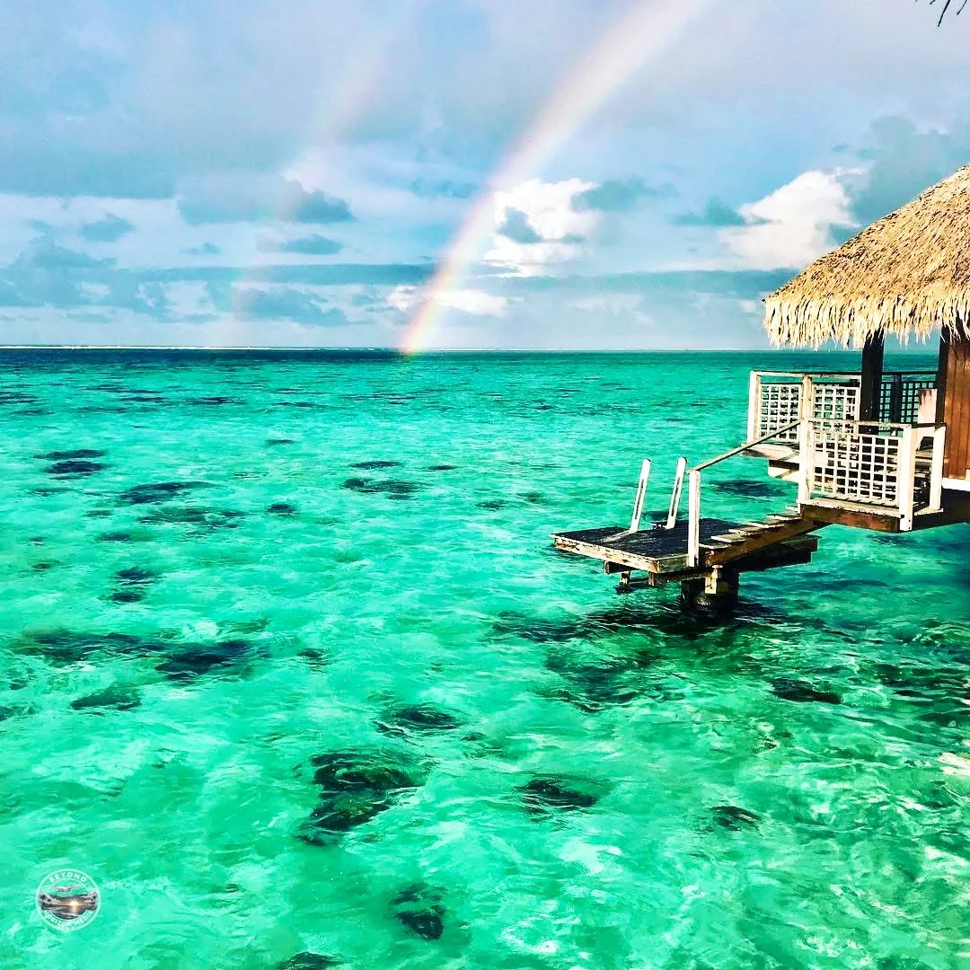 Overwater bungalow with a thatched roof, wooden deck, and stairs leading into clear turquoise water, with a rainbow in the cloudy sky in a tropical destination.