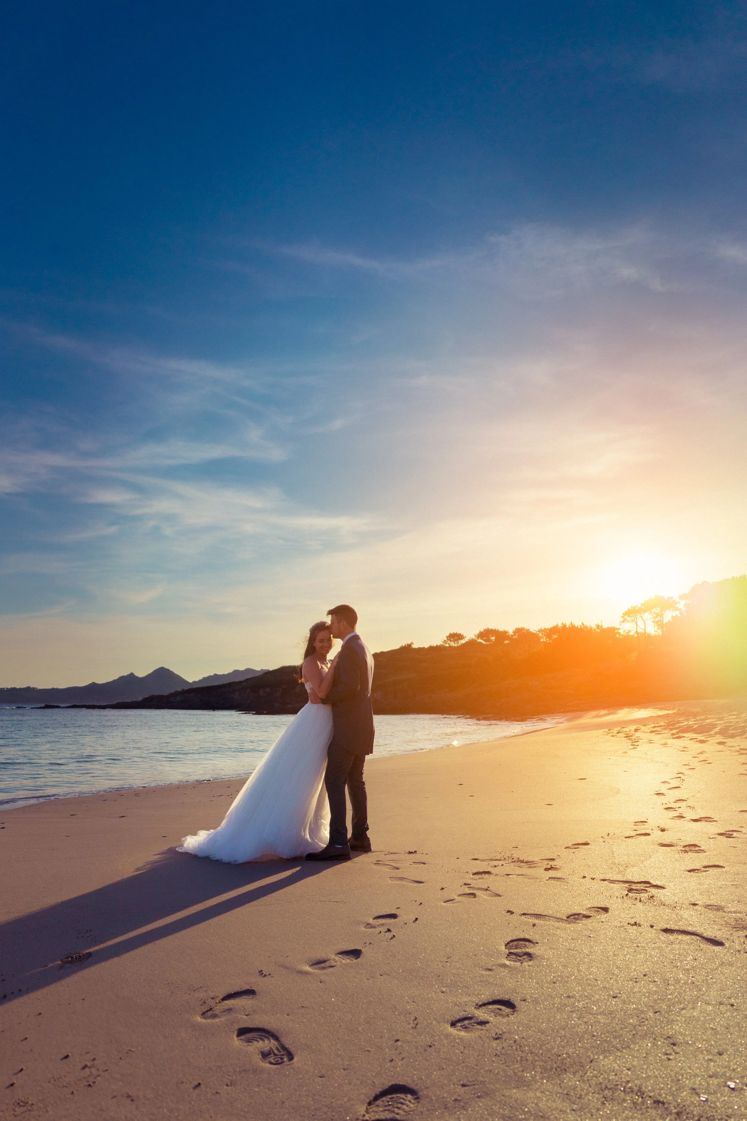 A bride and groom sharing a kiss on a beach at sunset with footprints in the sand.