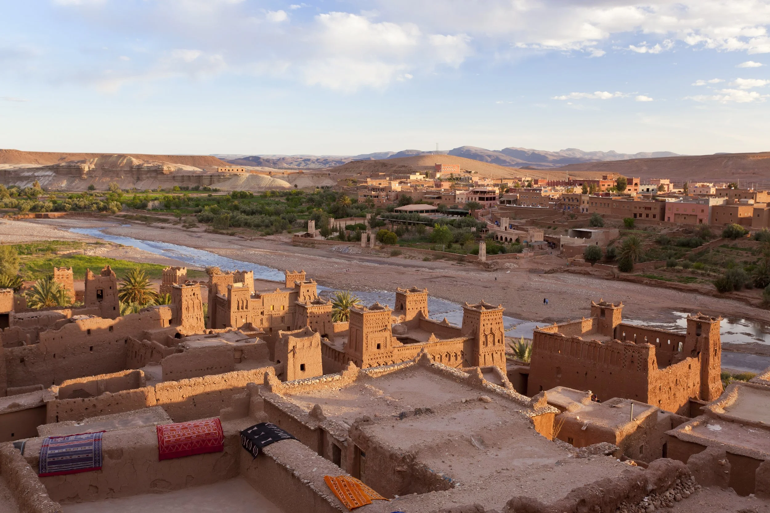 A desert landscape with traditional mud brick buildings, a river, palm trees, and distant hills during sunset.