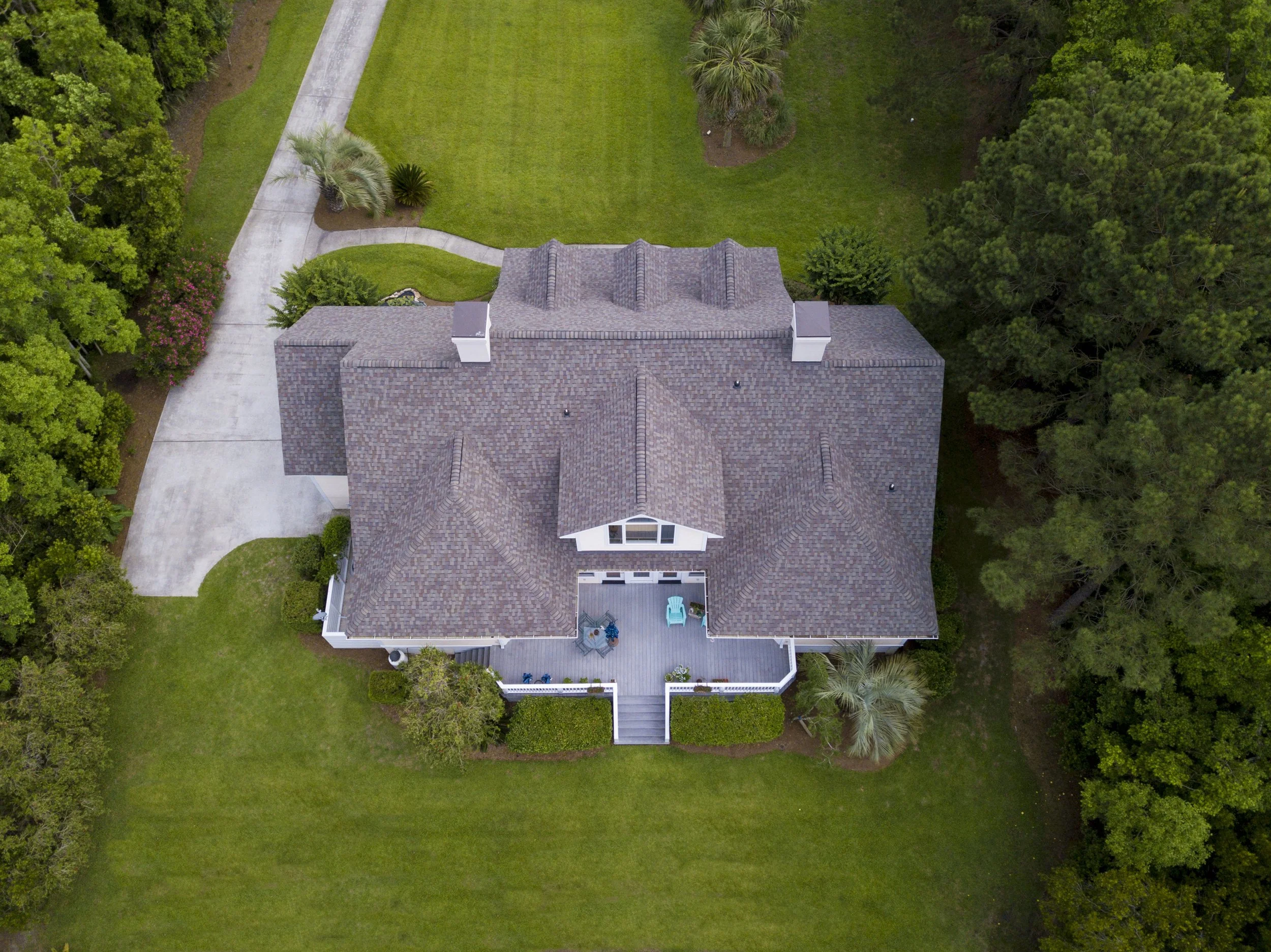 Aerial view of a large house with a gray shingle roof, surrounded by green lawn, trees, and a curved driveway leading to the house.