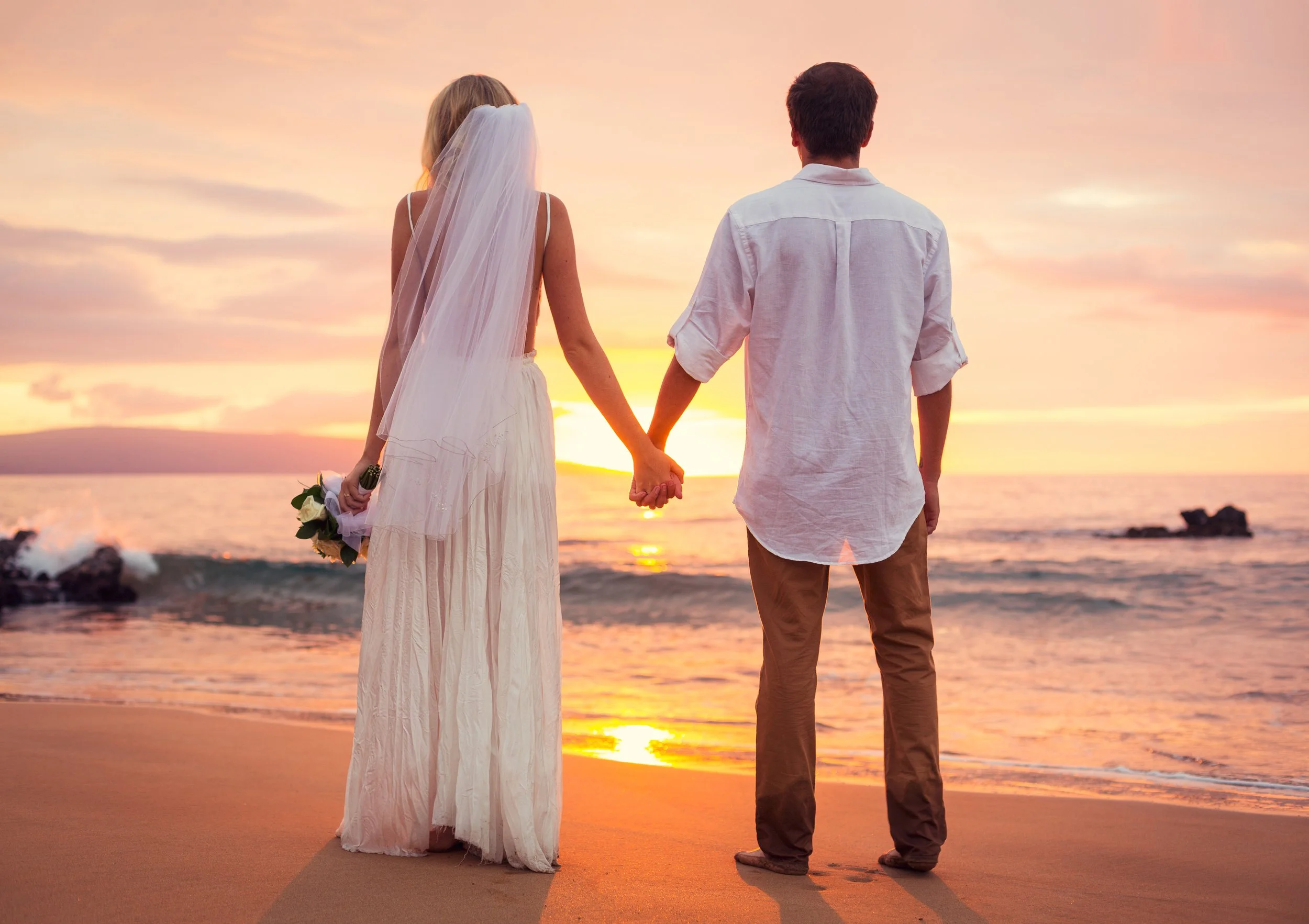 A couple, dressed in wedding attire, holding hands and facing the sunset on a beach.