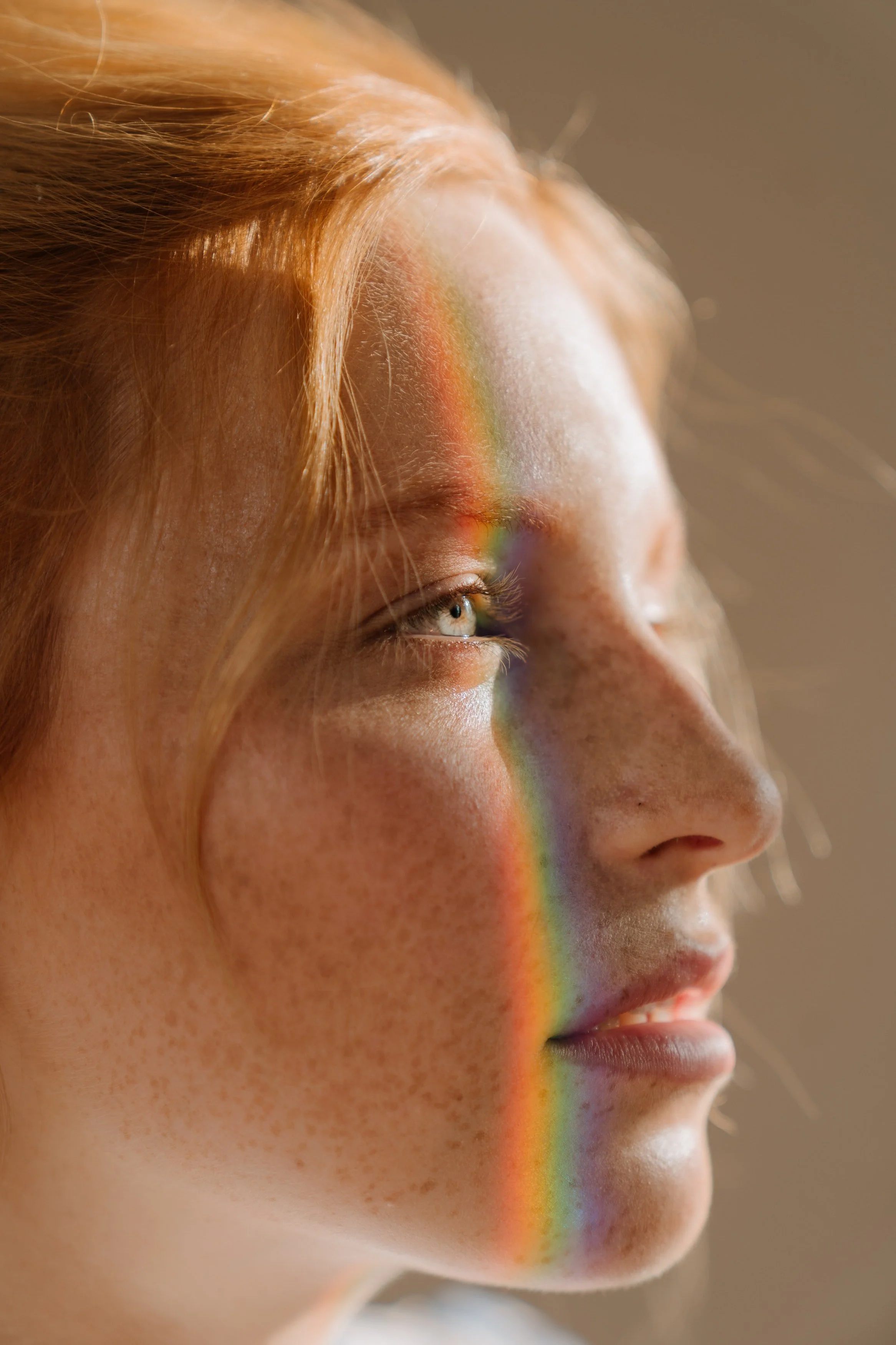 Close-up side profile of a woman with red hair, fair skin, freckles, and blue eyes. A rainbow pattern of light falls across her face from the left.
