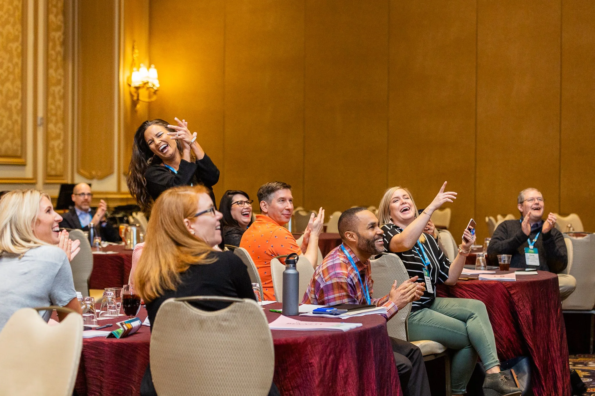 A diverse group of people attending a conference or event, sitting at tables, clapping and laughing, with some raising their hands in the air.